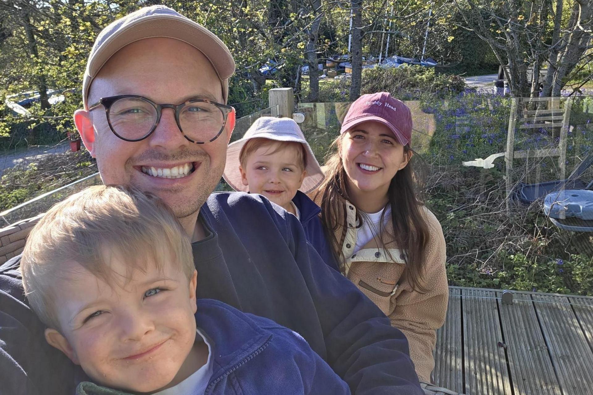 Kim Borthwick with her husband Ross and four-year-old twin sons Max and Freddie. She has long, dark hair, her sons have blond hair and blue eyes and her husband. She and her husband are wearing baseball caps and one of their sons is wearing a bucket hat. They are in an outdoor setting with trees behind them and are all smiling 