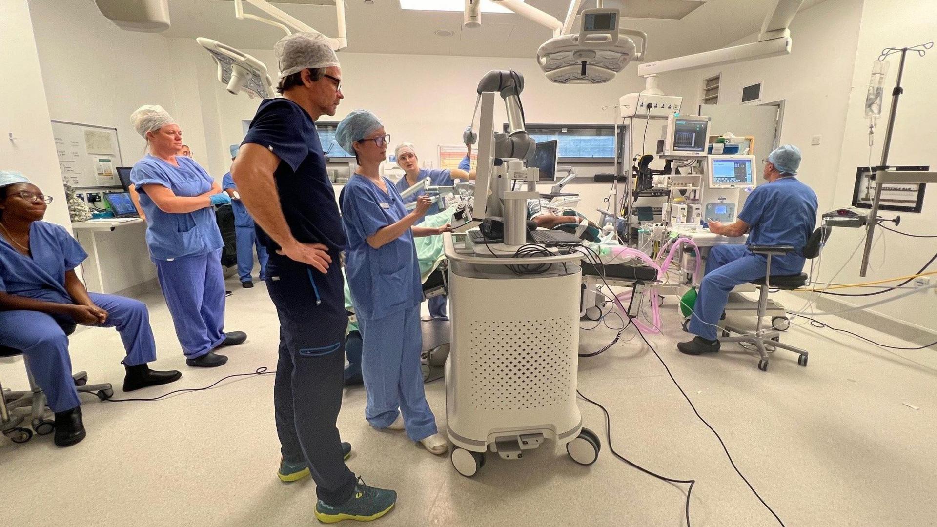 Several people dressed in blue medical scrubs are standing in an operating theatre looking at various machines and computers