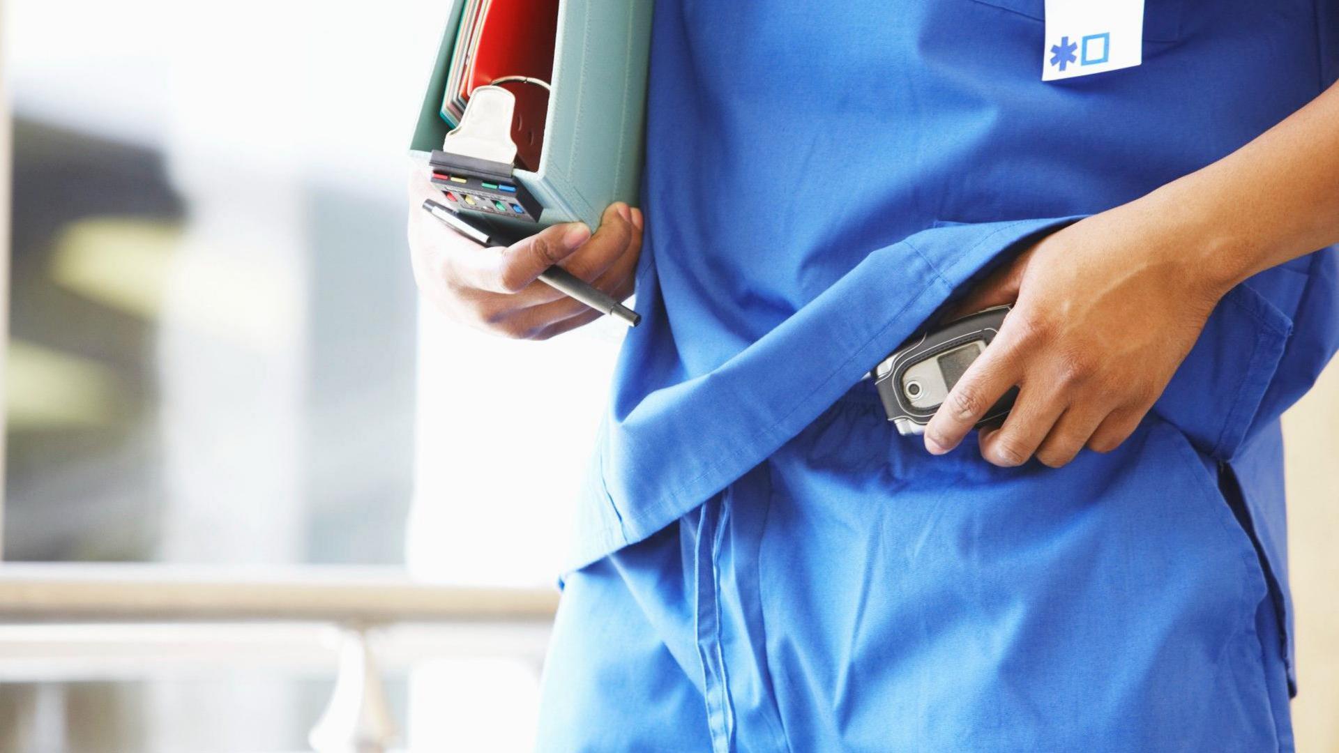 A doctor dressed in blue scrubs puts a pager on his trousers.