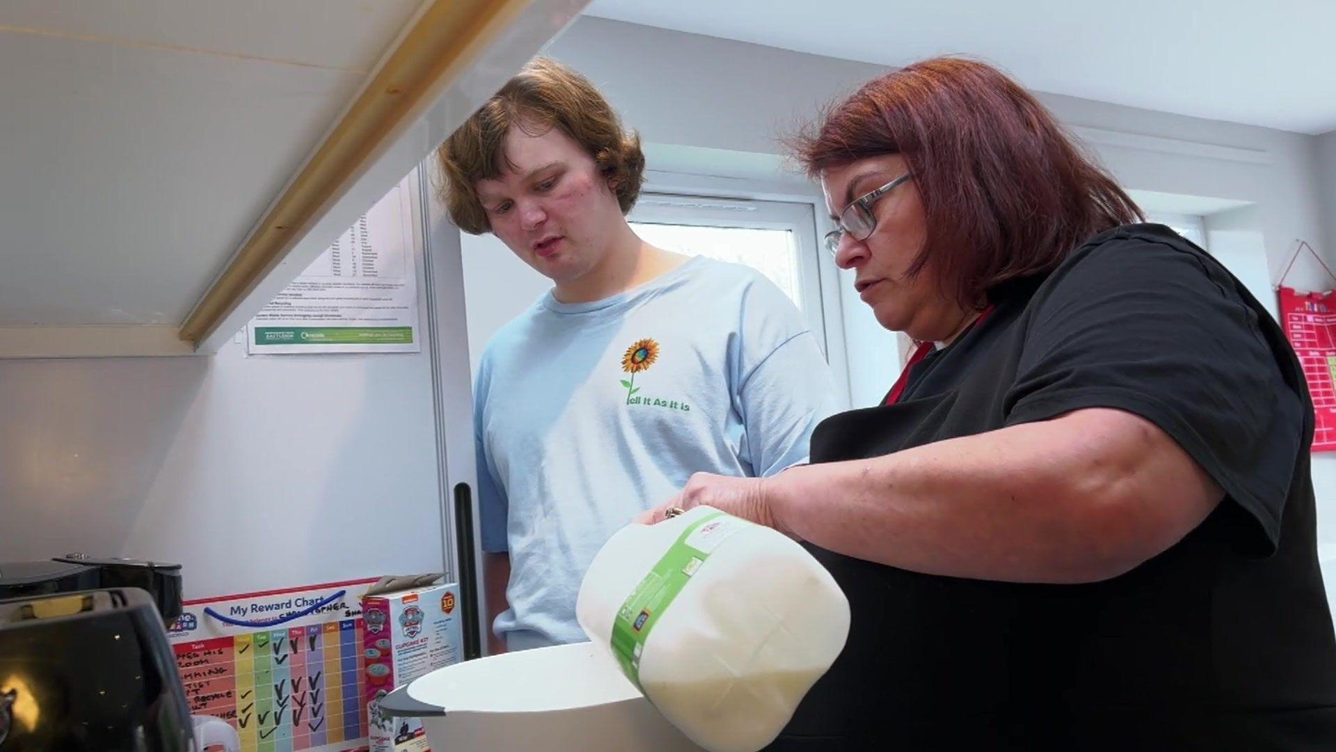 Christopher, with blonde hair wearing a blue T-shirt, watches as a woman in a black T-shirt pours milk into a mixing bowl. They are standing in a kitchen with a window behind them