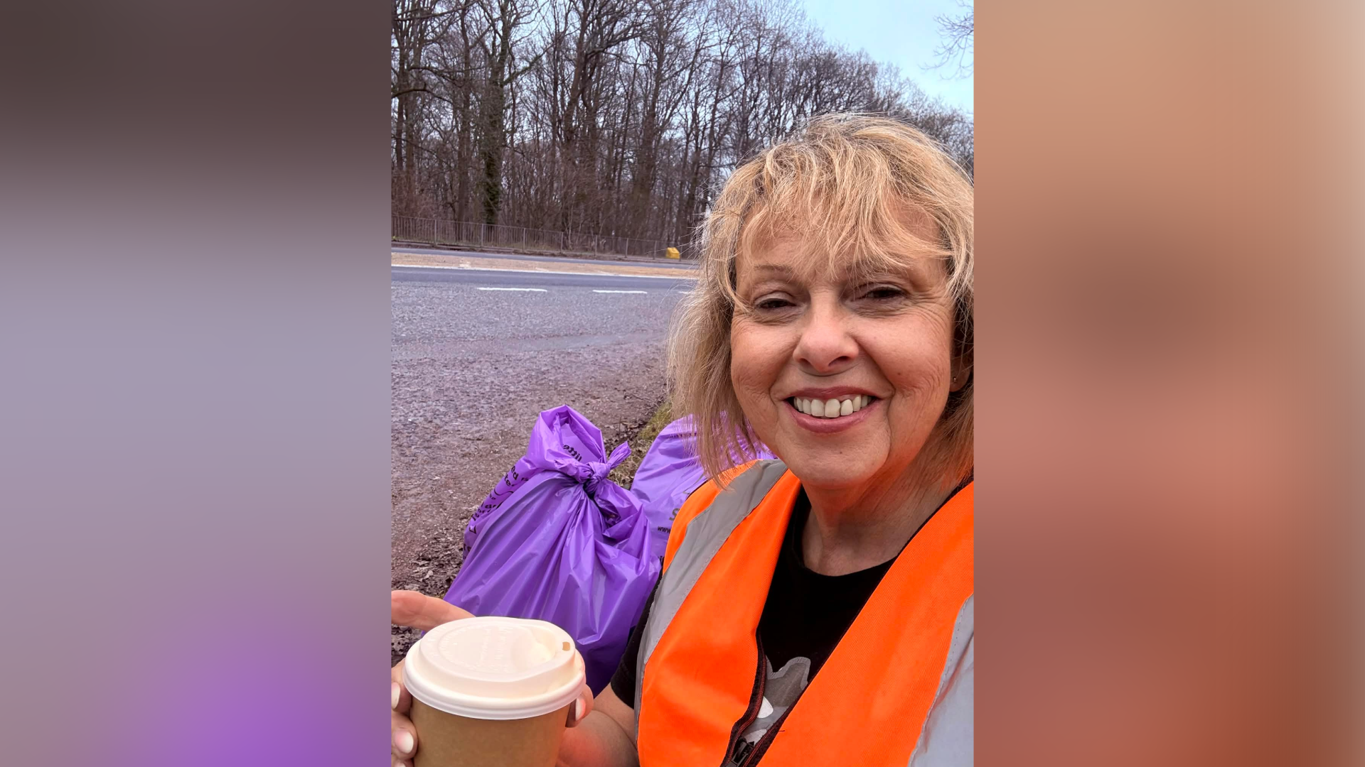 A woman with blonde shoulder-length hair is taking a selfie in an orange high viz jacket while holding a disposable coffee cup with purple plastic bags in the background