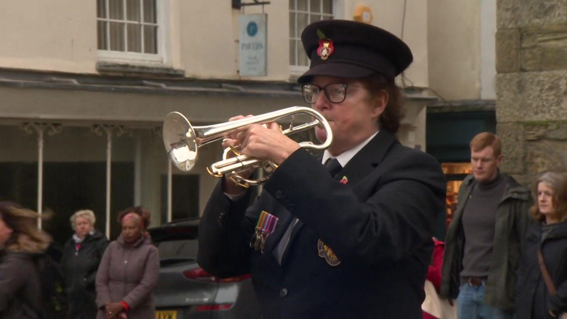 A woman is dressed in her uniform and playing the bugle to mark the beginning of the two minutes silence. Several people are stood behind her.