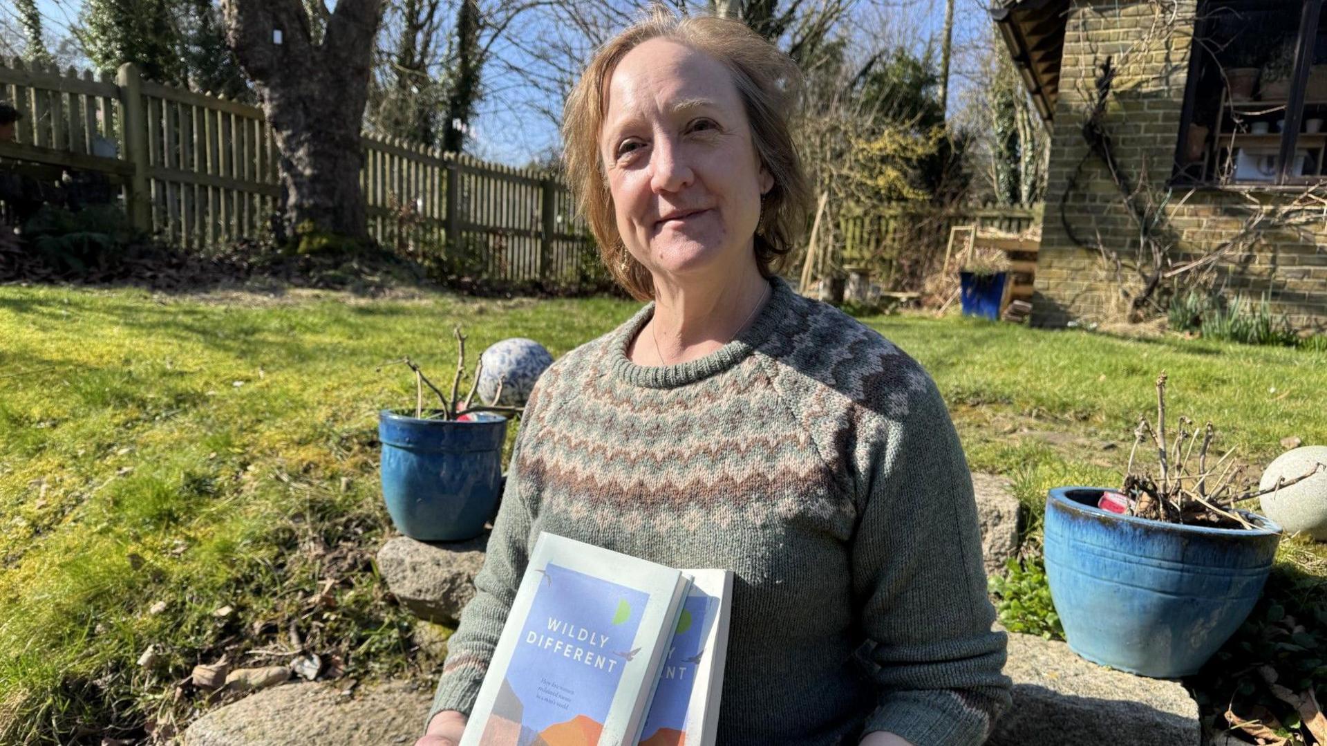 A woman sat in a garden holding a book titled "Wildly Different"