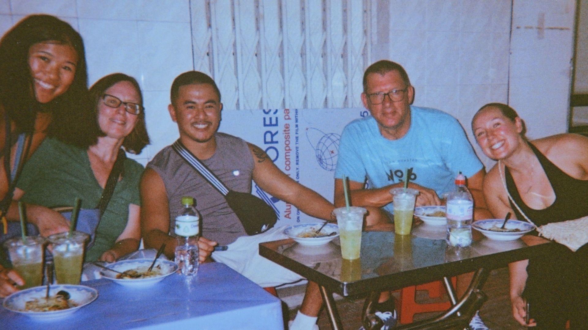 Ike and his adoptive family are all sitting on smiling around a table. Ike is between his mum and dad. His sister and girlfriend are also smiling to the camera.
On the table there are bowls of food and plastic cups with green straws.