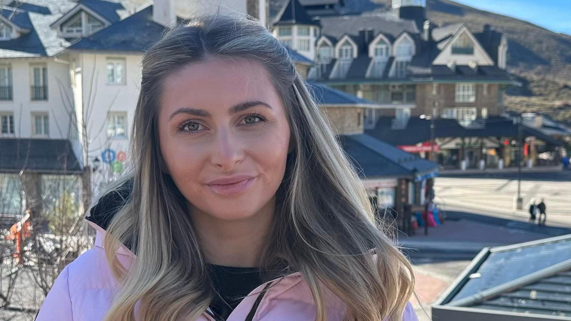 Amber Mason, standing outside, looking straight at the camera. She has long hair, that is tied back at the front, and down. She is smiling, and there are buildings behind her. 