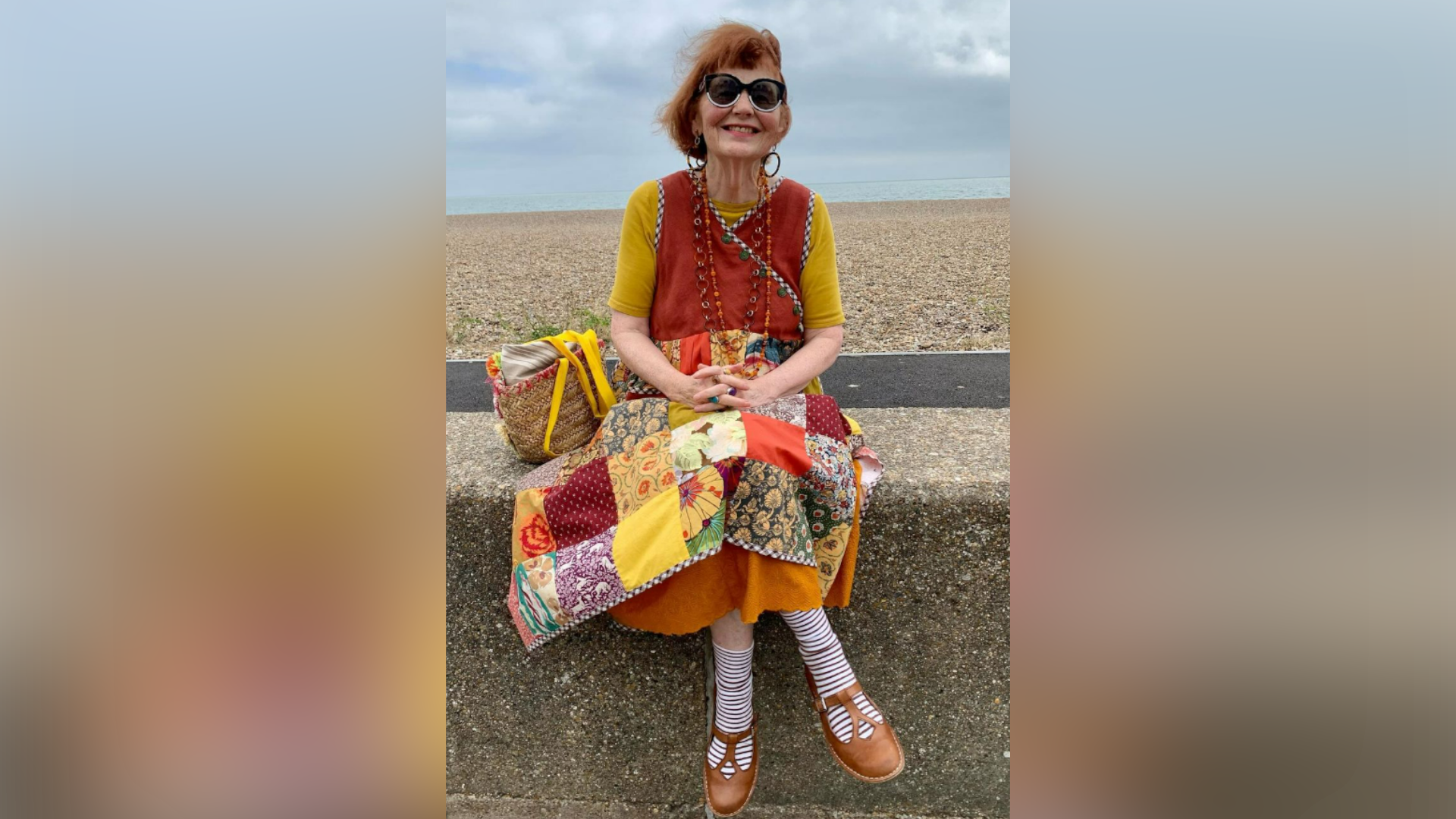 Marcia Riddington with red hair sits on a sea wall smiling at the camera. She wearing a yellow top with a red vest over the top and a skirt made up of a former quilt with different patches of fabric.