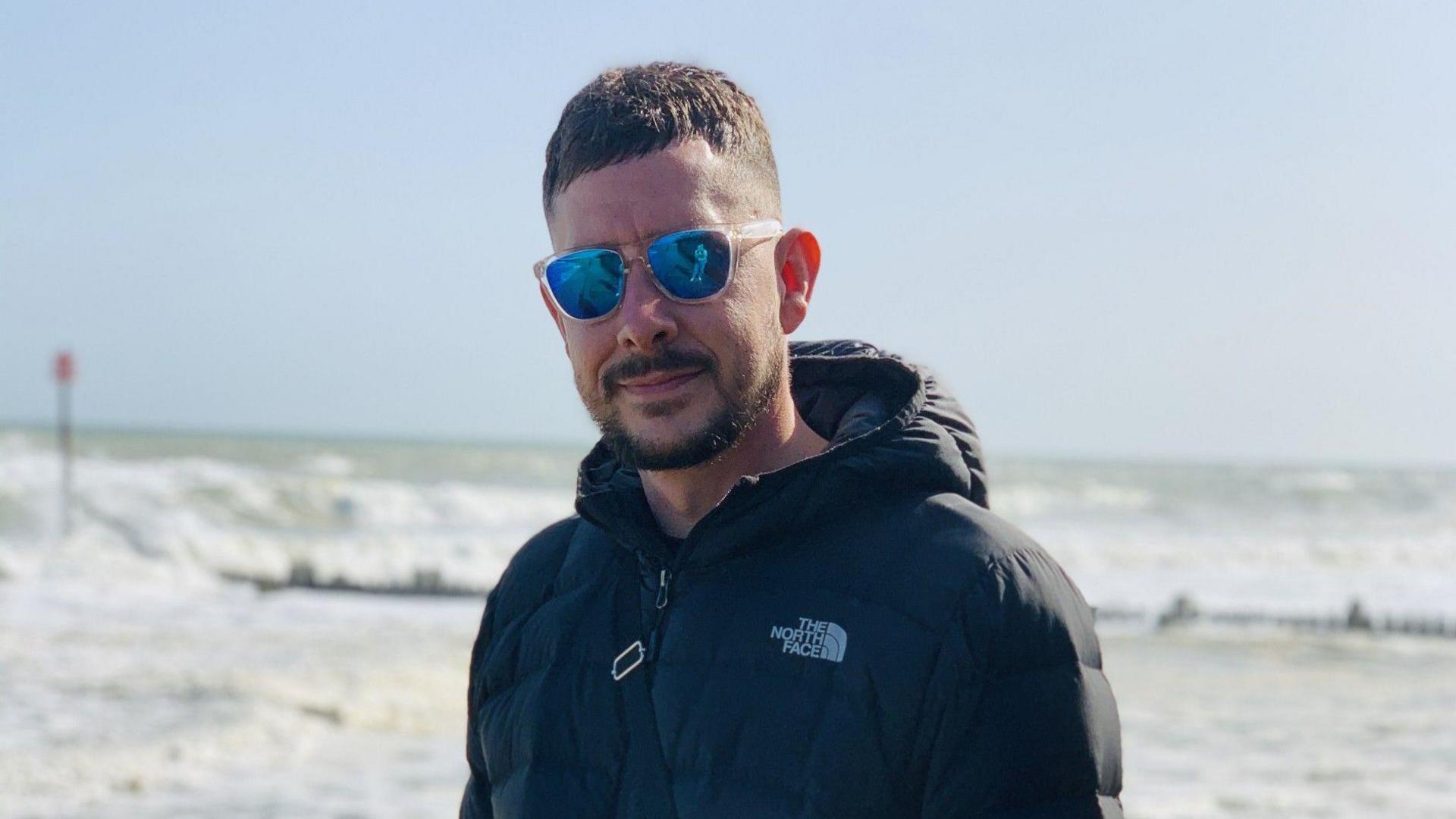 A man with short brown hair and short beard and moustache standing by the sea. He is wearing sunglasses and a padded jacket.
