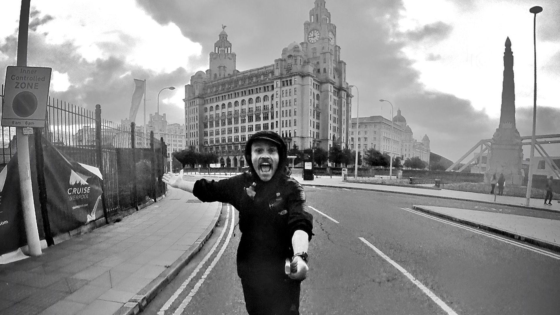 Andy Hobson, running along a city centre road in Liverpool. The photo is in black and white, with the city's Three Graces visible in the background. 