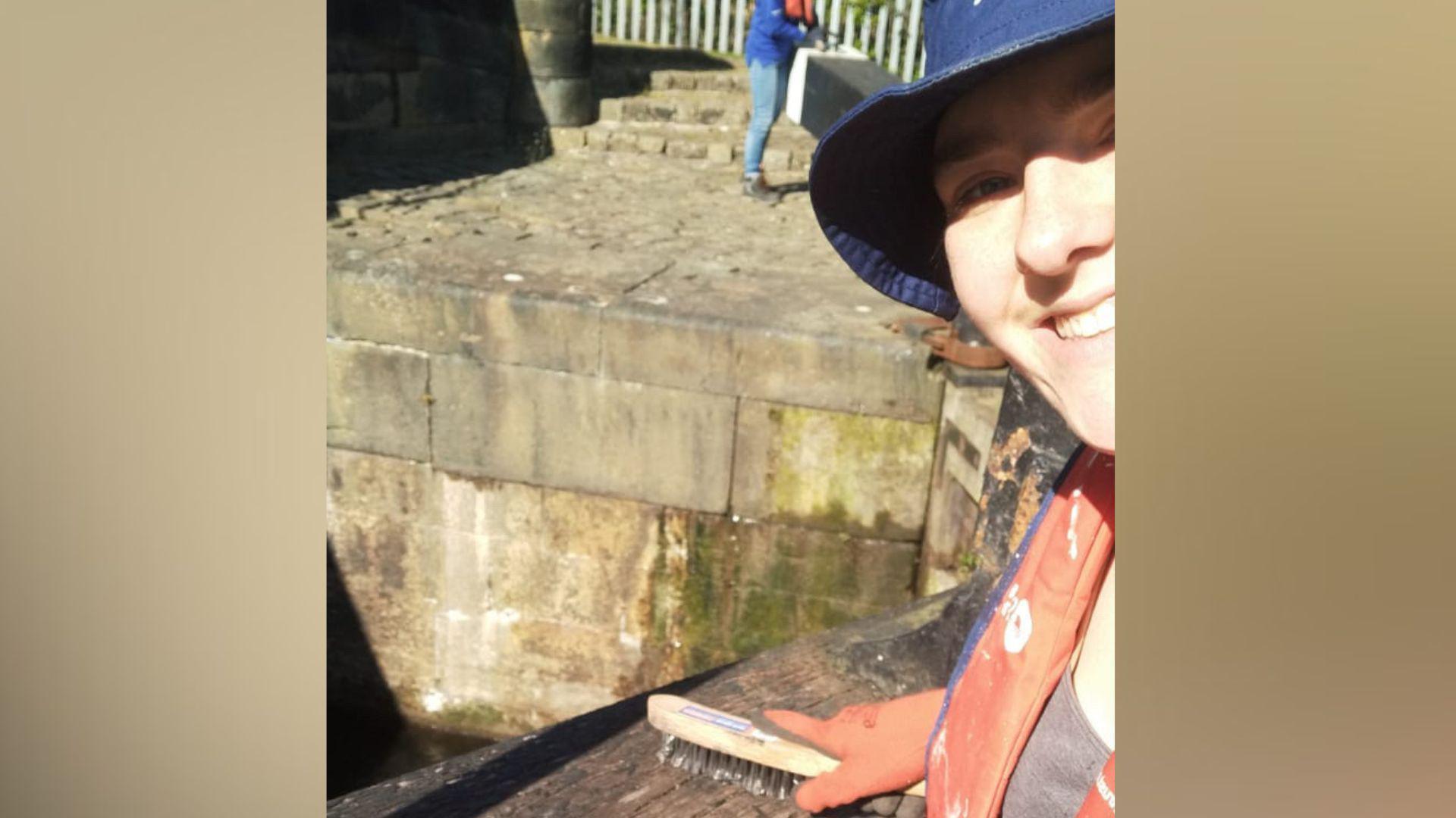 Eve is wearing an orange life jacket and blue clothing and holding a scrubbing brush against a dark wooden surface near a stone canal lock. The background shows a stone-paved area with moss on the walls and a metal fence, with another individual standing further away.