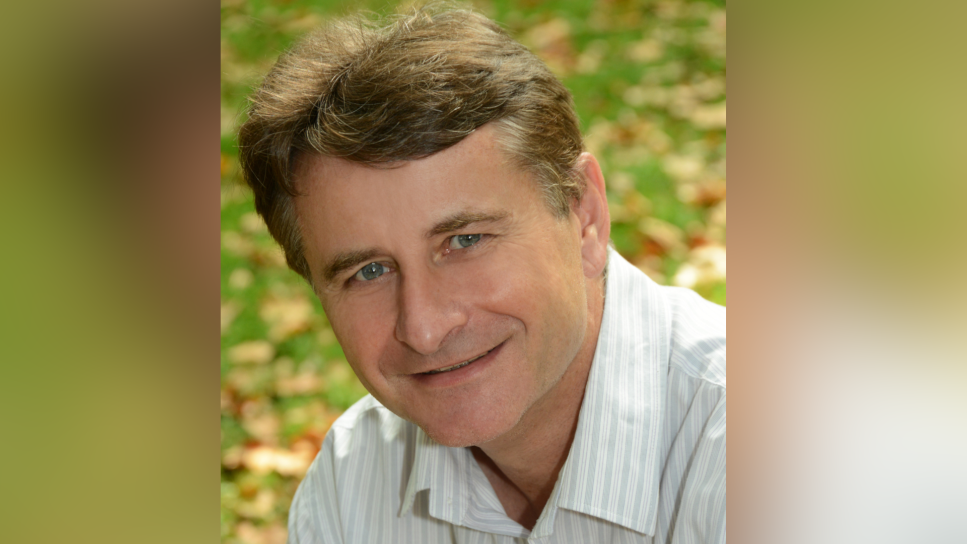 A man looks into the camera. He has blue eyes and short brown hair. His head is tilted and he is wearing a striped shirt. Behind him are orange and brown leaves on some grass. 