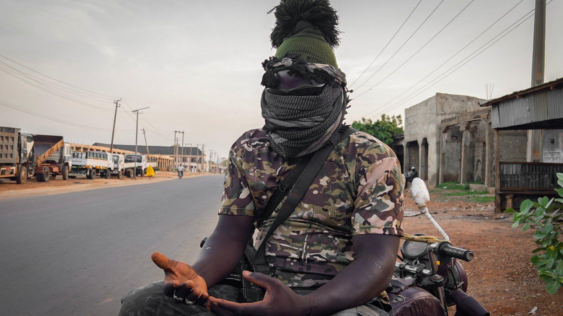 A antheral wearing a camouflage-patterned top, his look covered with scarves, sits connected a motorbike, a weapon successful his thigh and his hands unfastened successful beforehand of him, palms up