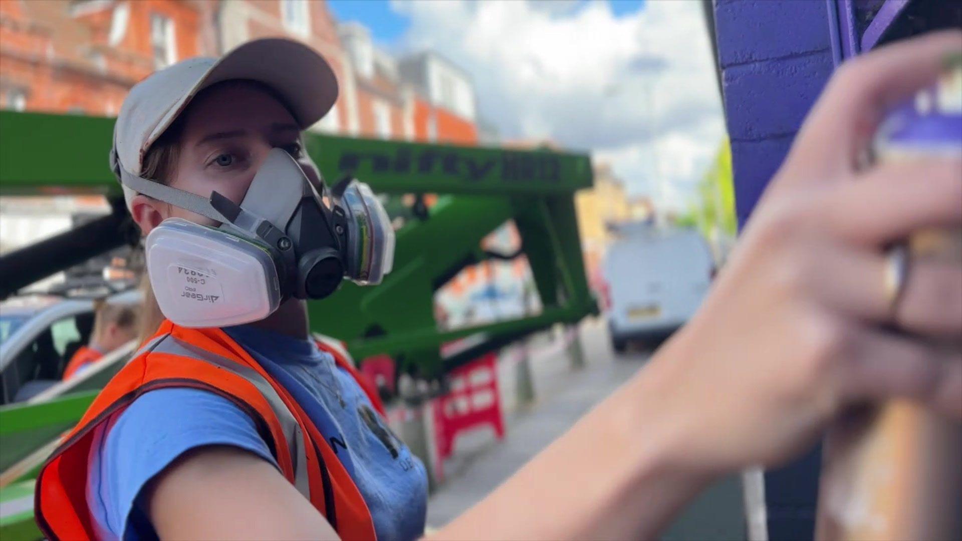 A female artist is pictured spray painting a wall in Ipswich. She is wearing a pink cap with a mask over her face to protect her from the paint fumes. She also wears a blue top with an orange high-vis vest over the top.