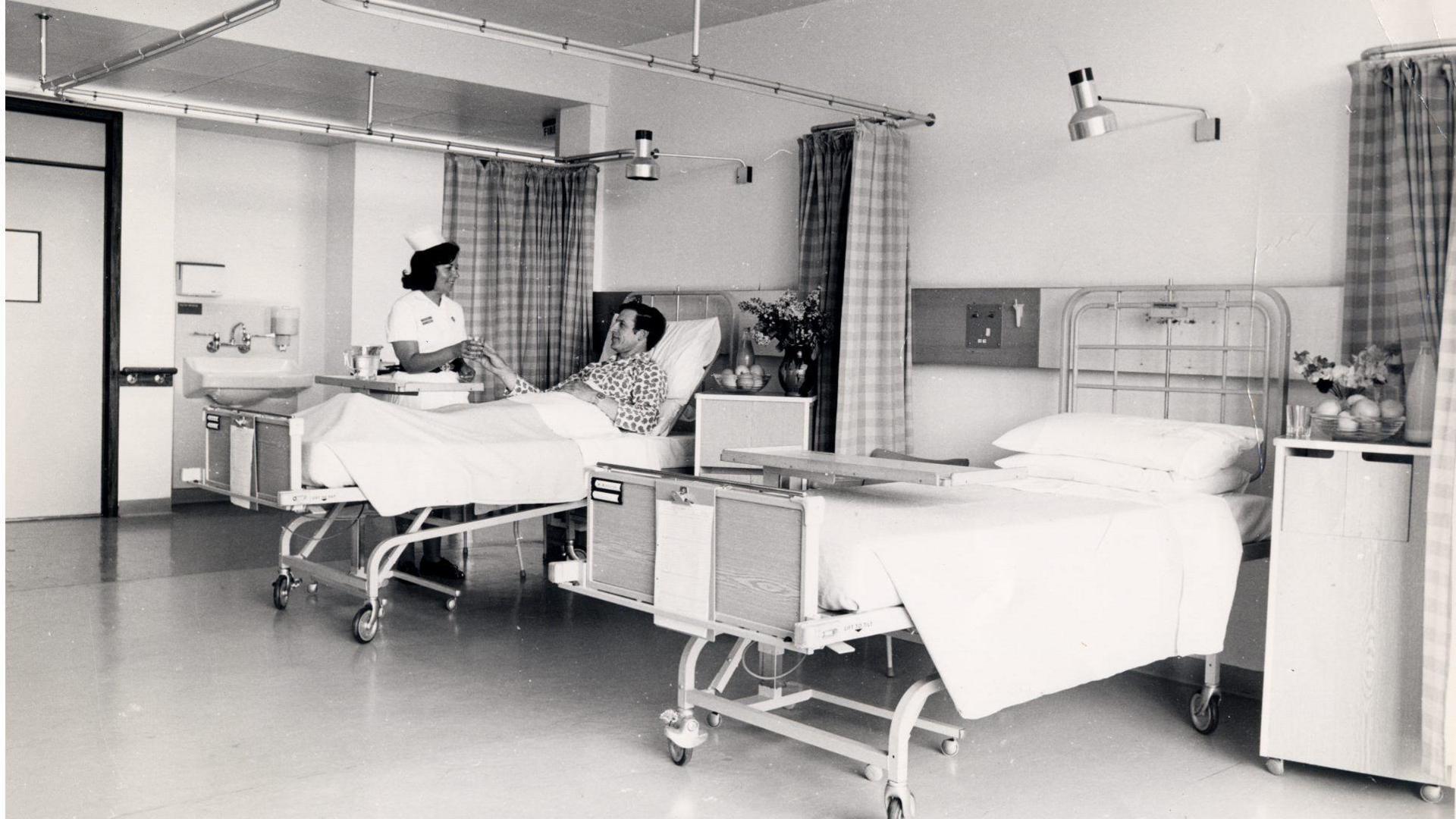 Two beds sit in a hospital ward in this black and white photo. In one, a man in spotted pyjamas looks up to a hospital nurse.