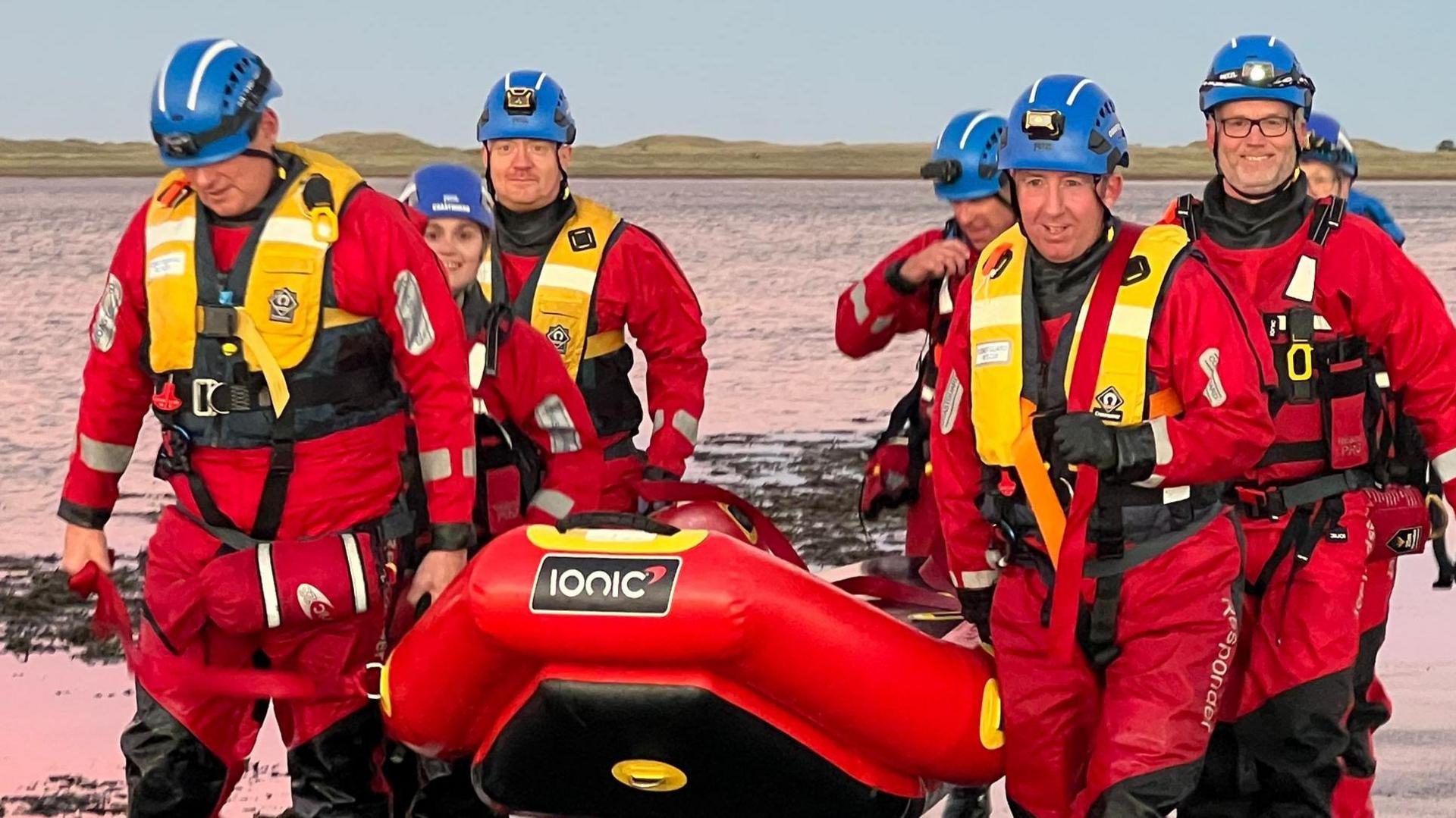 A group of seven rescue officers wearing red dry suits, yellow life jackets and blue helmets. They're carrying a red and yellow inflatable sled. The sea can be seen in the background.