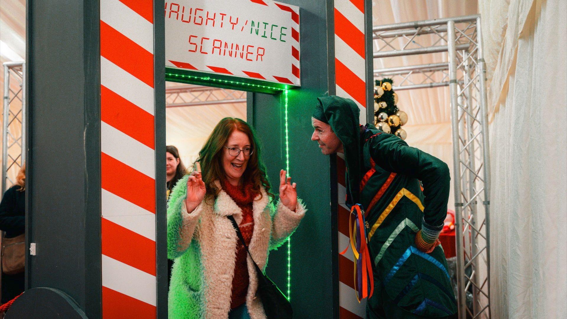 A man dressed as an elf looking at a women who has her fingers crossed as she goes through a body scanner, with the words 'naughty or nice' above it. 