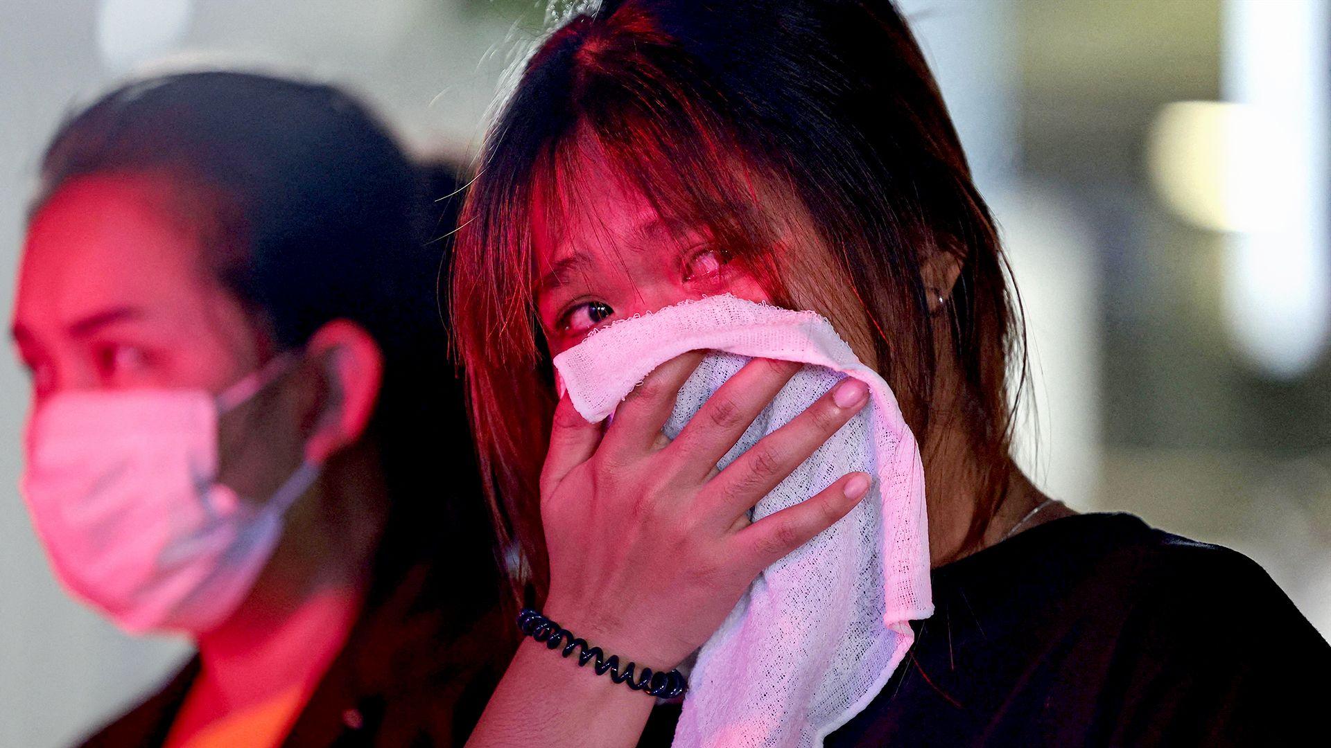Two women stand, their faces lit by emergency services lights, one wearing a face mask and the other holding a white cloth over her mouth and nose. 