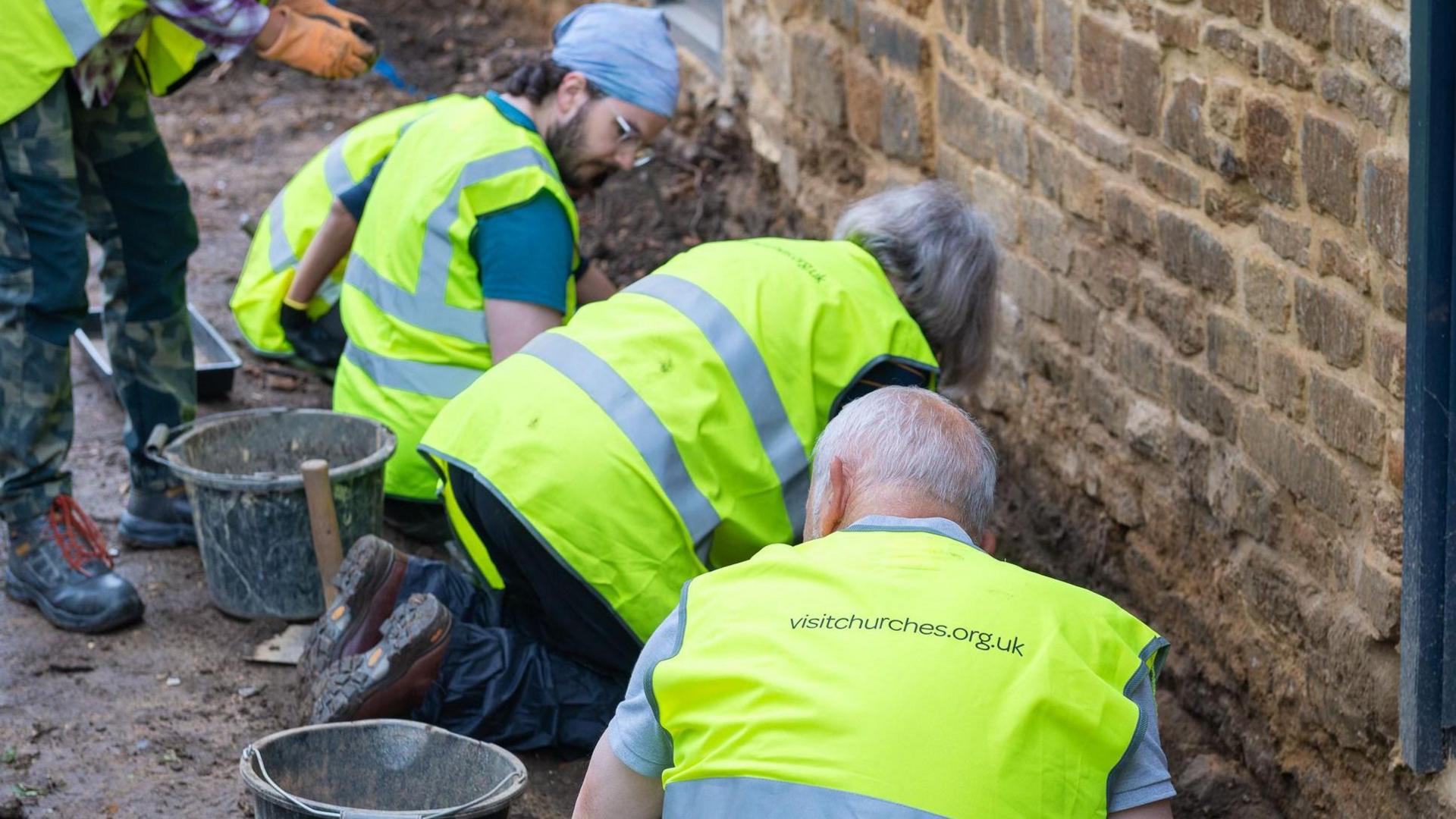 People in yellow hi-vis jackets kneeling against brick wall with metal buckets