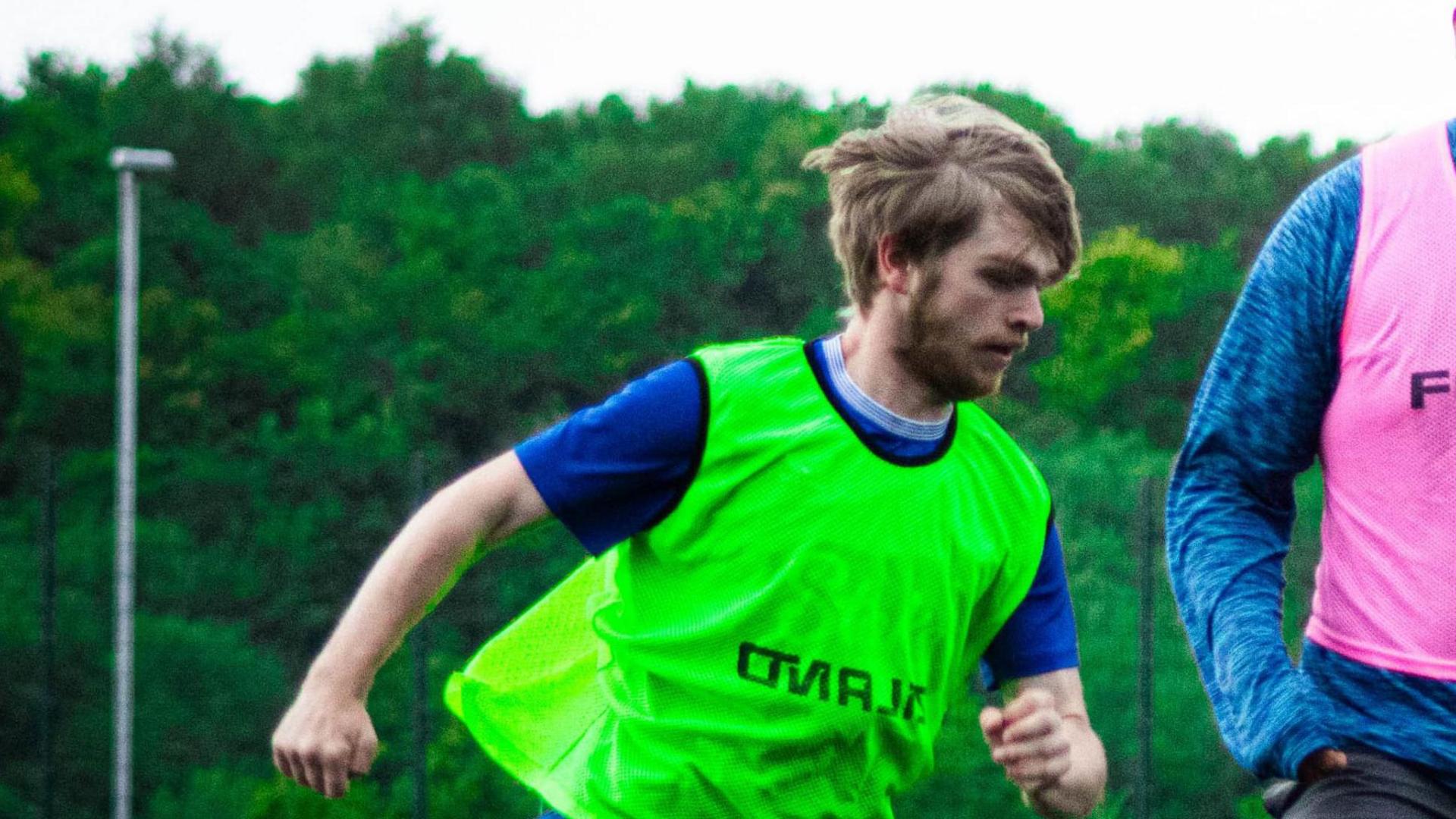 Ray Ferguson with long blond hair and beard playing football wearing a green football bib and blue t-shirt.