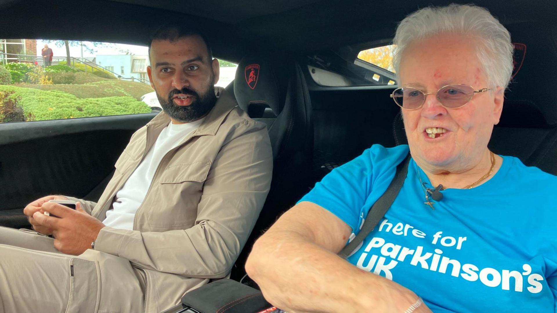 A smiling woman wearing a blue T-shirt which says 'Parkinson's UK' on. She is sat as a passenger in a car next to a man who has a black beard and is wearing light beige trousers, a jacket and white T-shirt.