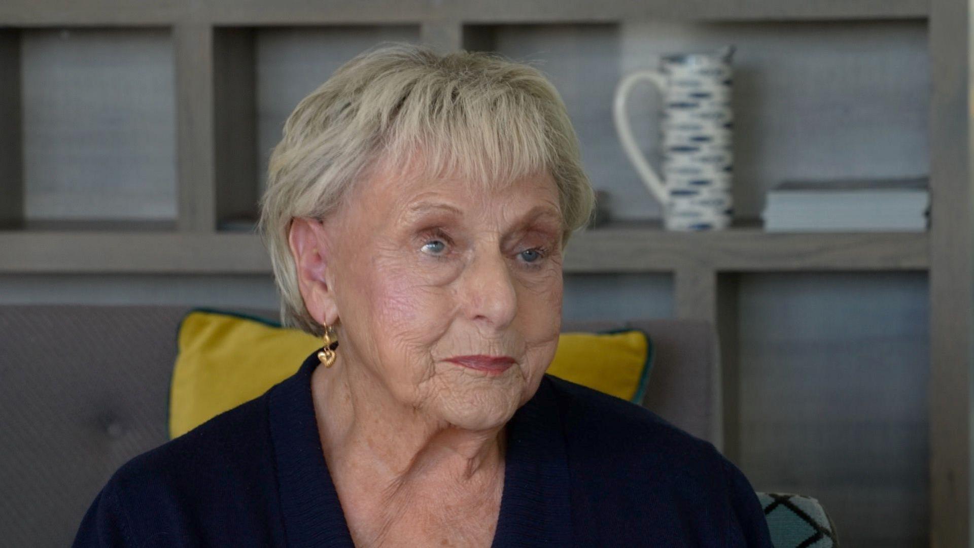 Ann Ming looking to the right of the camera. She has short blonde hair and is wearing gold heart earrings. She is also wearing a black top and is sitting in front of some grey shelves.