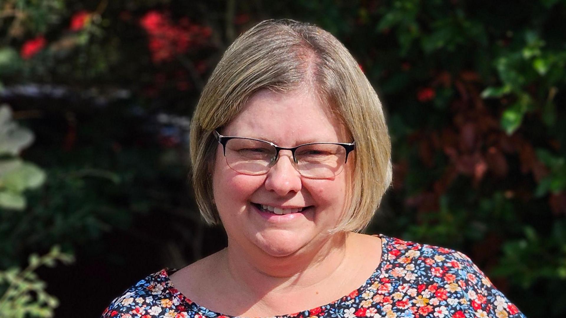 Jo is wearing a floral dress and spectacles and is stood in front of a bush