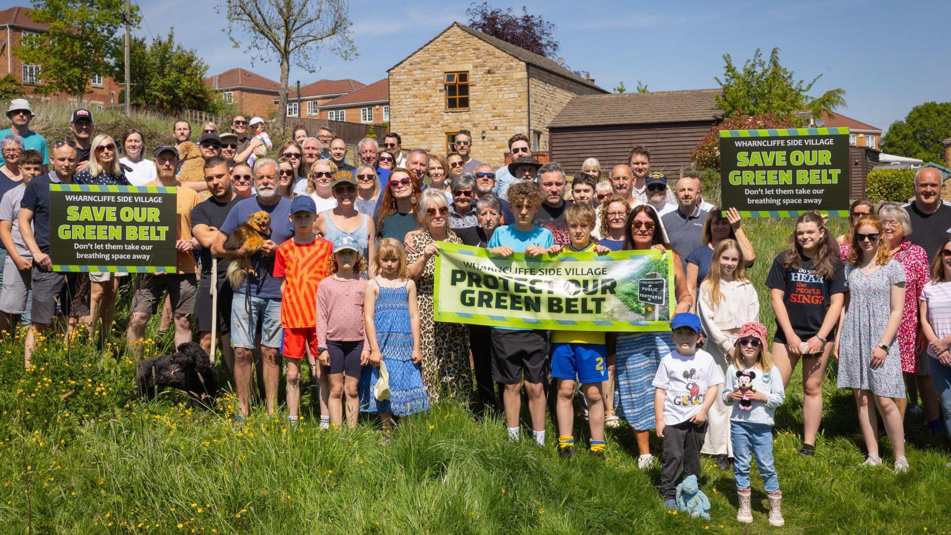 Dozens of people are stood in a green field, with houses in the background. They are holding placards and banners saying "protect our green belt"