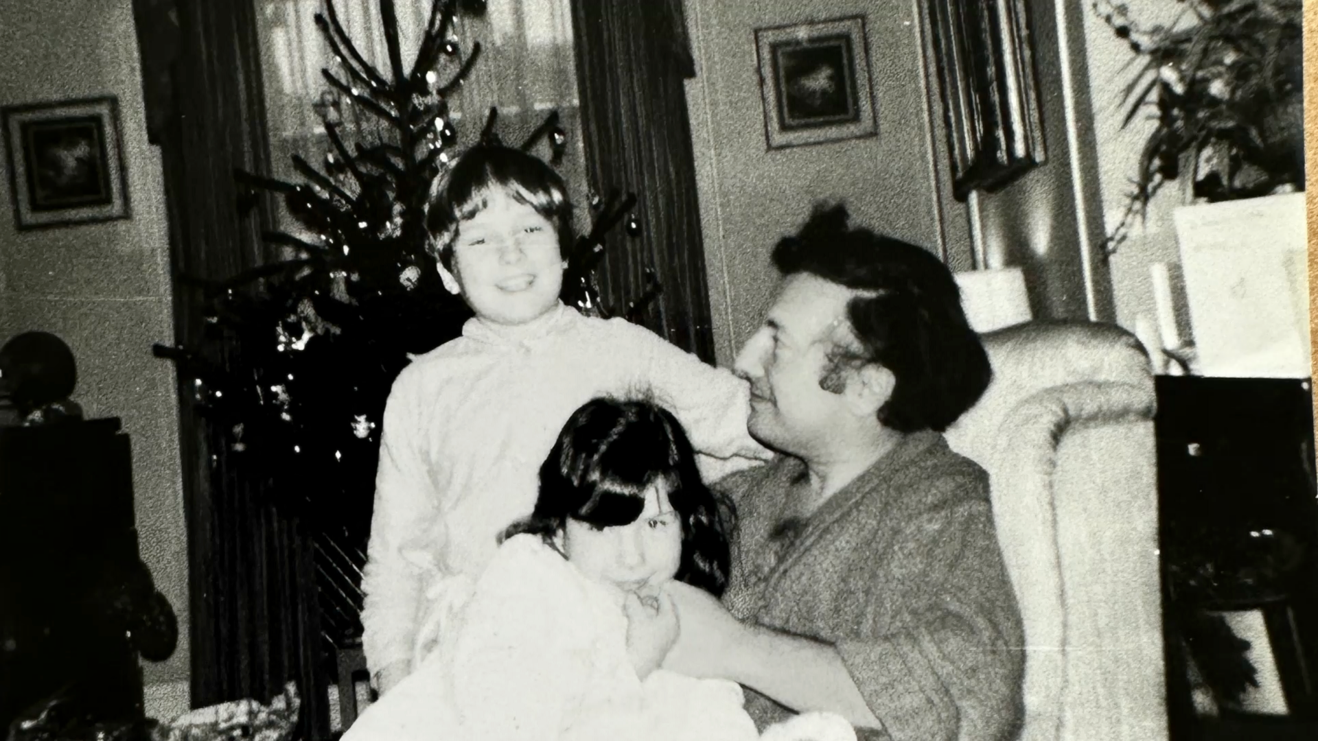 A black and white photo showing a young boy and girl sitting on their father's knee in an armchair in front of a Christmas tree. The three are smiling.