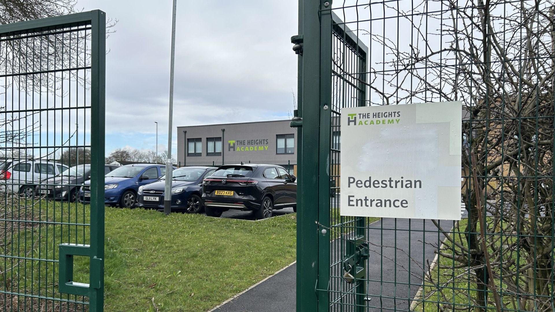 The Heights Academy in Smith's Wood. A pedestrian entrance sign hangs on a green metal gate at an entrance to the building. The school building is in the background and a car park can also be seen. 