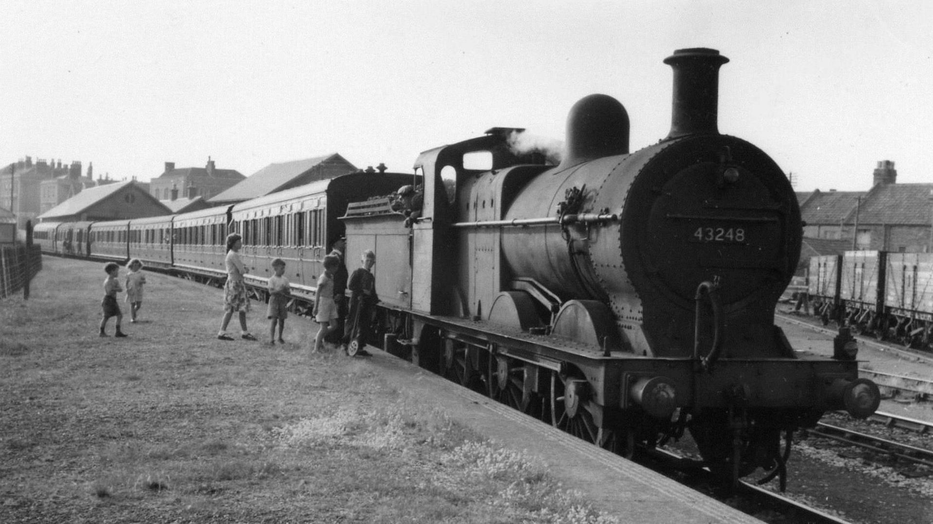 A group of children in the 1950s talking to engine driver of a large black steam locomotive. The image is black and white and slightly overexposed.