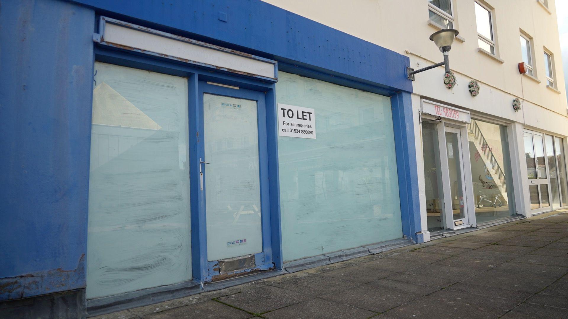 An empty retail site with a blue shop front. There is a to let sign on the shop window. Next to the shop is two other vacant retail sites. 