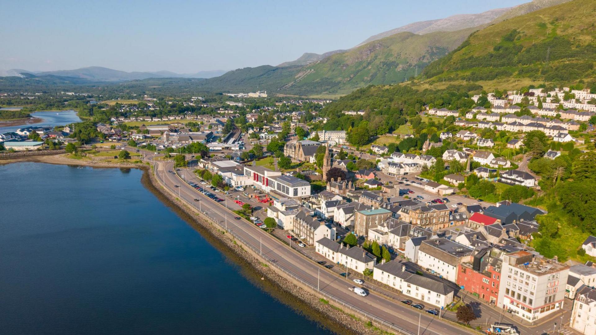 An aerial view of Fort William on a sunny day. The image shows the A82 where it runs along a shore of Loch Linnhe, and the rows of buildings that line the road and other properties built on a hillside overlooking the loch.