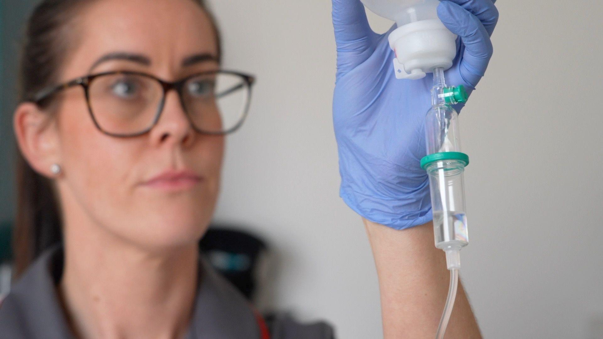 Matron Lana Goodwin holds up an intravenous drug drip. She has blue surgical rubber gloves on. Ms Goodwin is wearing brown glasses, has her hair tied back and is wearing a grey uniform with red seams.