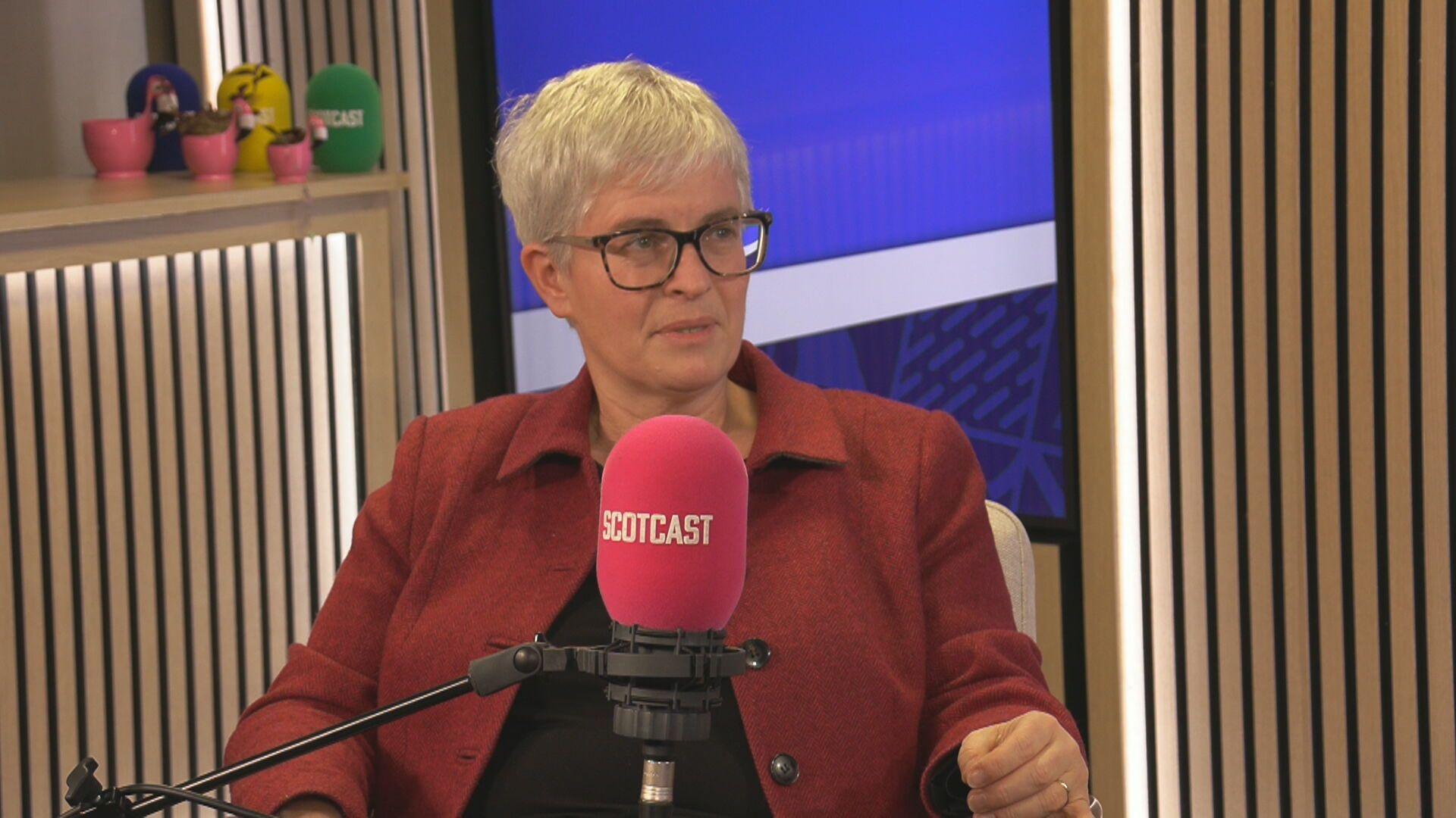Naomi Cunningham sitting in a BBC podcast studio. She is behind a microphone with a red cover that says Scotcast. She has short, grey hair and glasses. She is wearing a red jacket