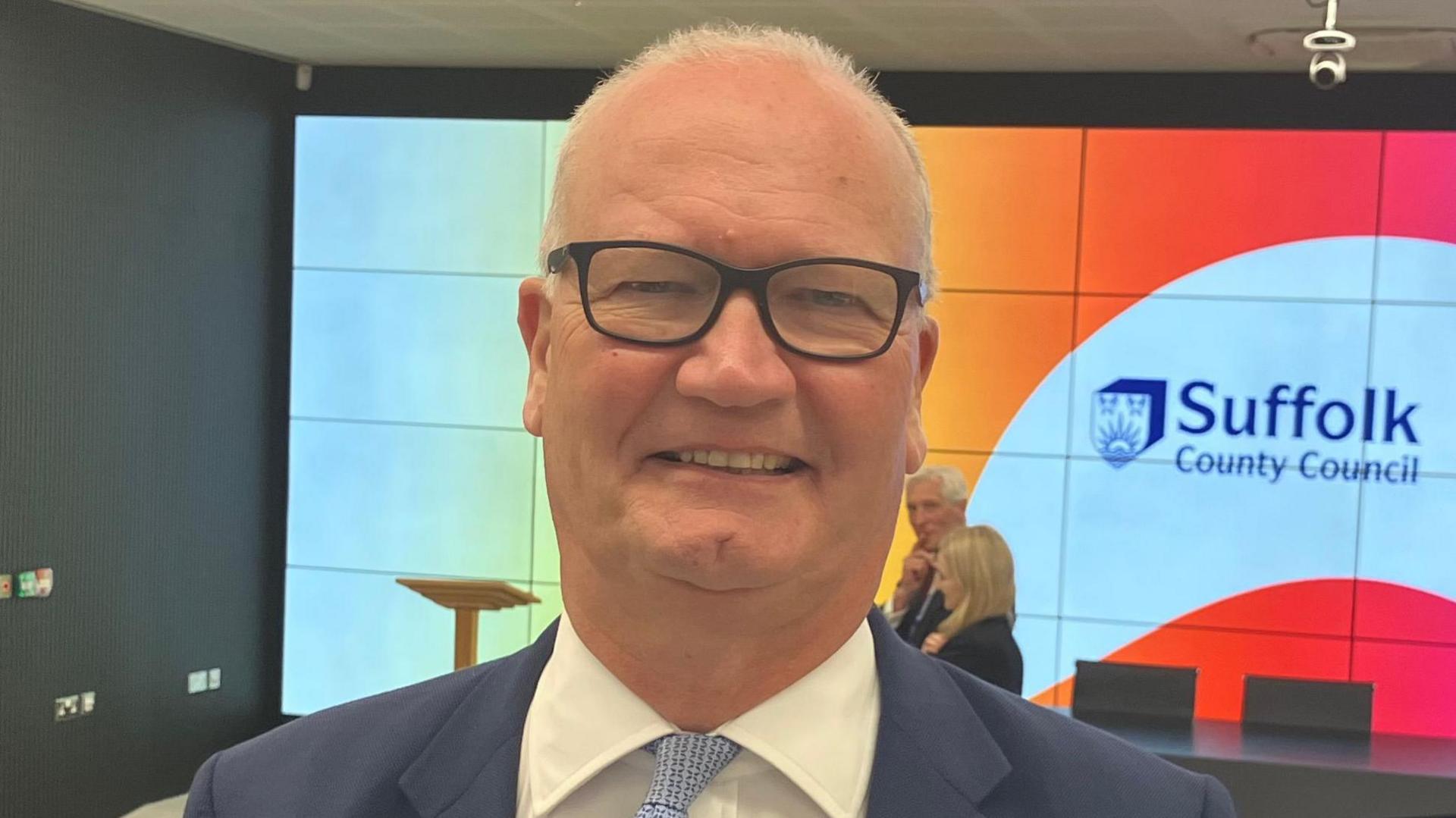Matthew Hicks looking towards the camera, smiling. He is wearing black glasses and a blue suit jacket with a white shirt and pale blue tie.