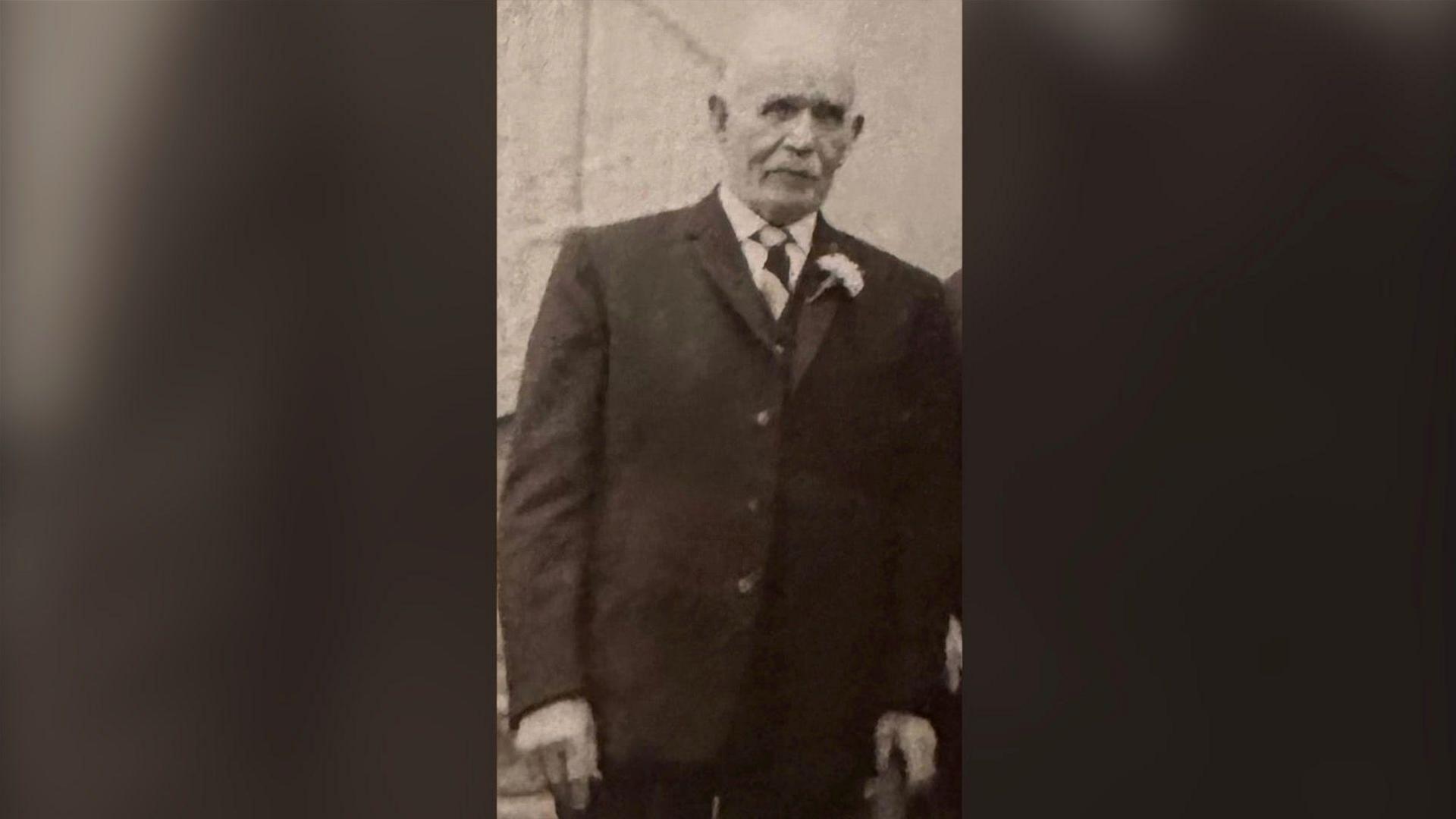 A black and white archive photo of the late Owen Pyers attending a wedding.  He is an elderly man with white hair.  He is wearing a dark suit, white shirt, a striped tie and has a flower in his buttonhole.