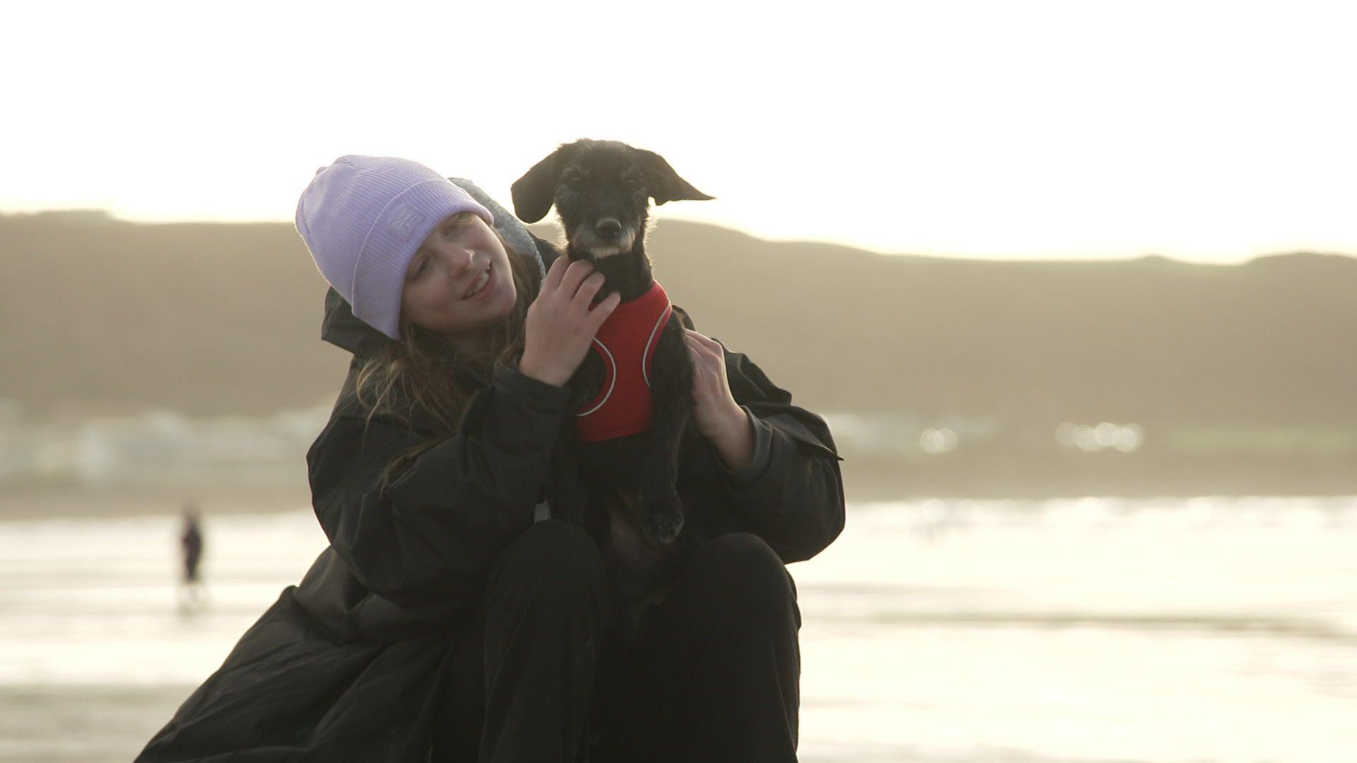A young girl sat on the beach with a purple hat and black coat on, smiling at her black and white dog who is sat on her knees wearing a red harness.