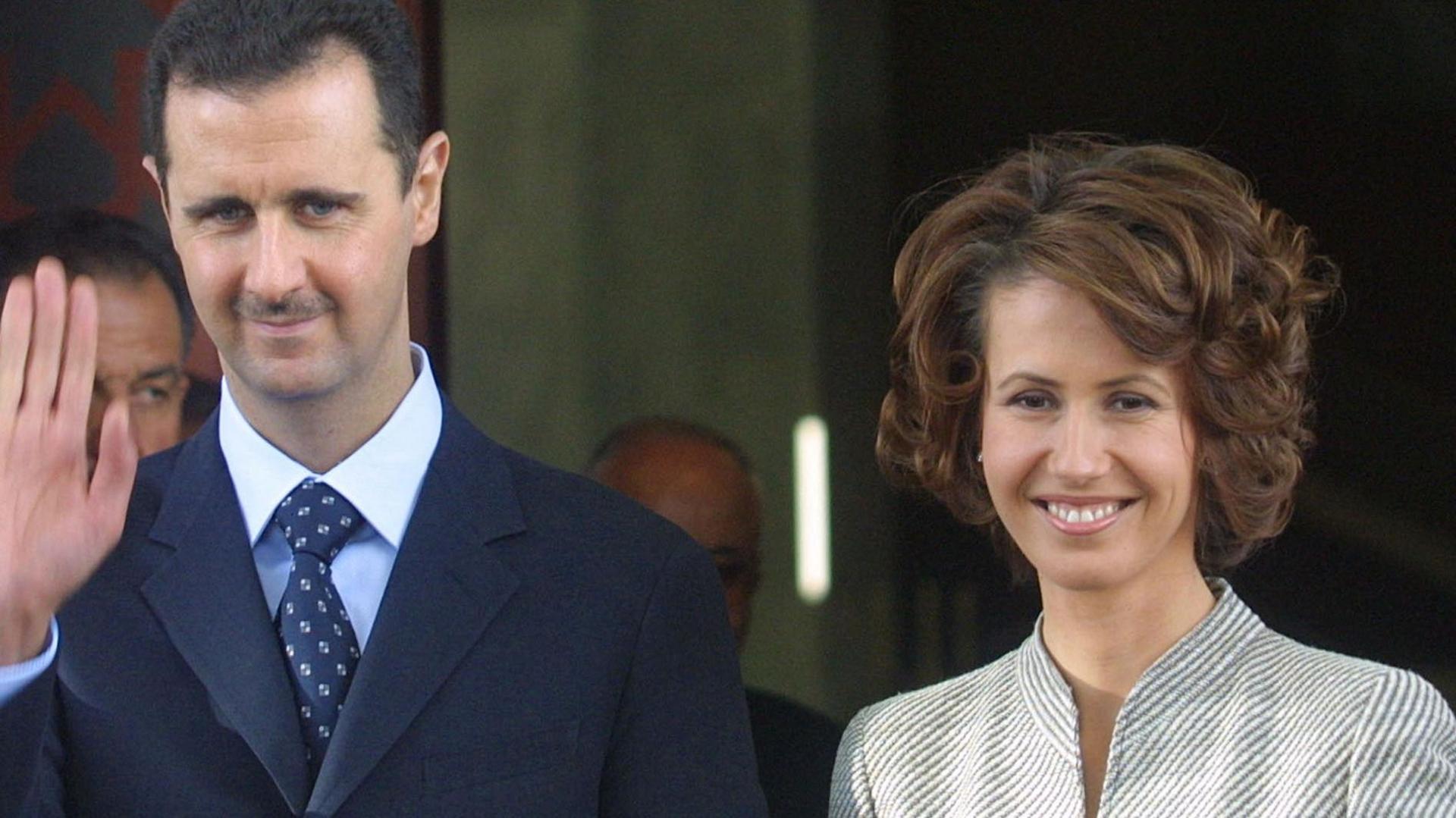 Bashar al-Assad, left, smiles and waves wearing a dark suit. His wife Asma al-Assad smiles more widely beside him, and wears a grey striped jacket.