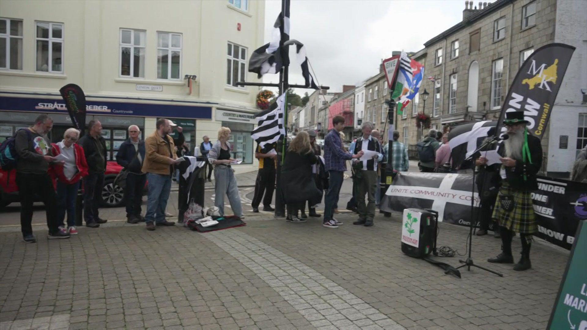 People gathered in Truro holding Cornish flags and looking towards the performer on the right. To the left of the performer is a large black and white Cornish flag. Behind the people gathered together is a white building.