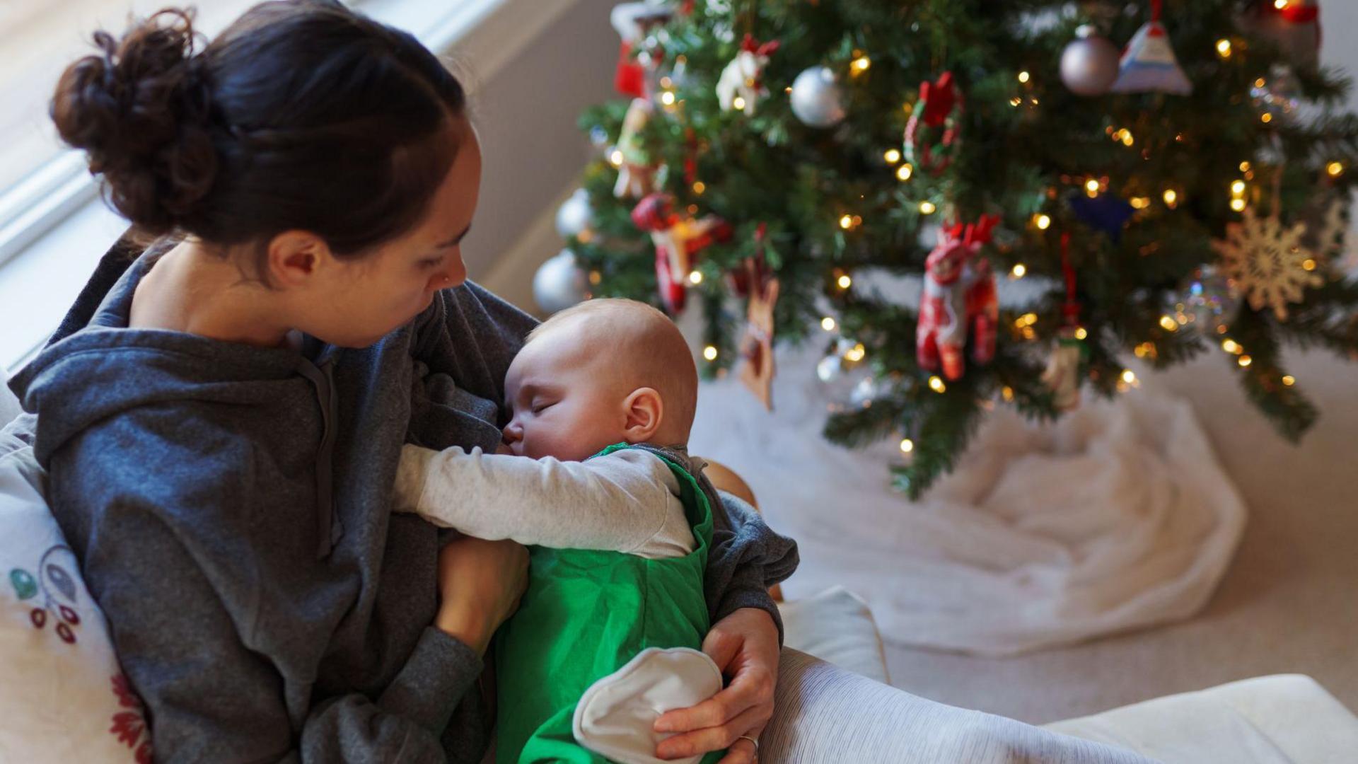 A woman sits on a sofa next to a Christmas tree and breastfeeds her baby