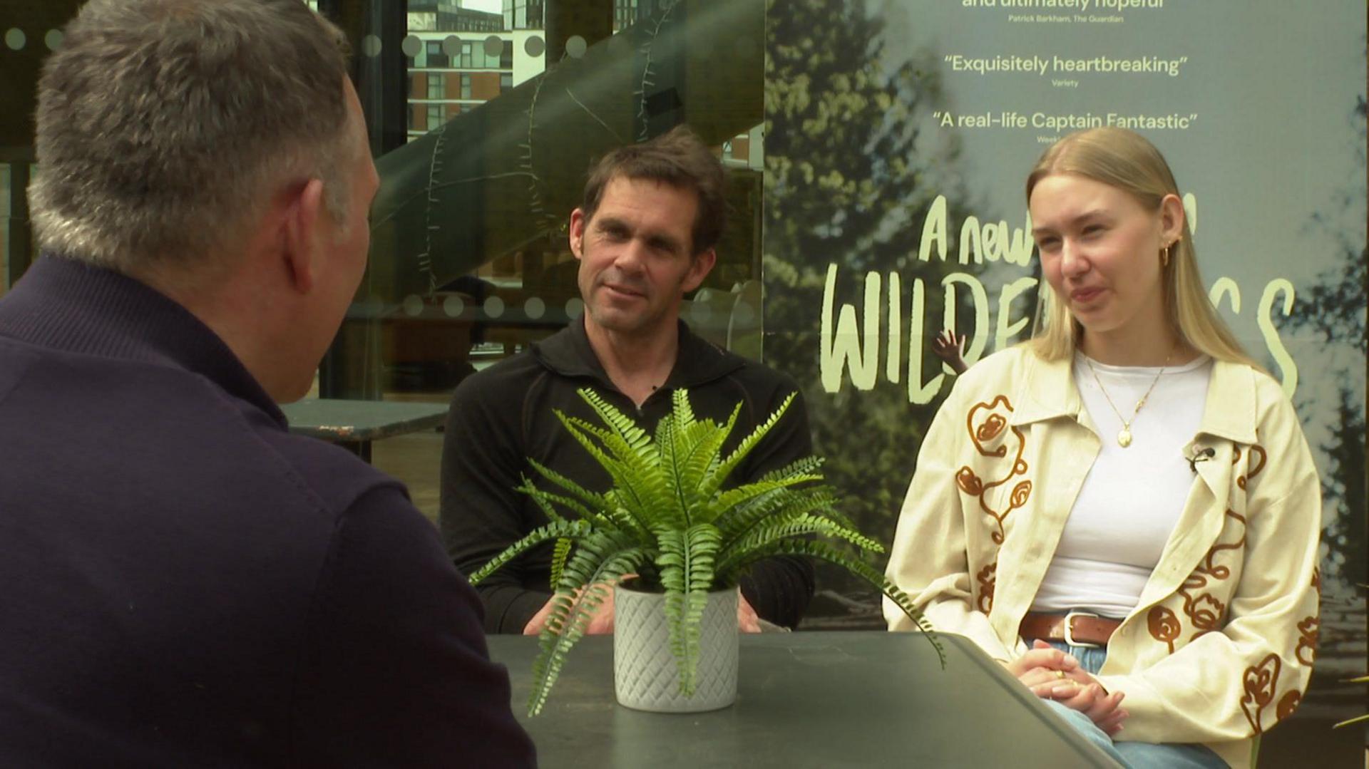 Nik Payne, who has dark hair and is wearing a black open-collared shirt, and Ronja, who is blonde and wears a cream overshirt over a white top, sit in front of a poster for the film and speak to the camera