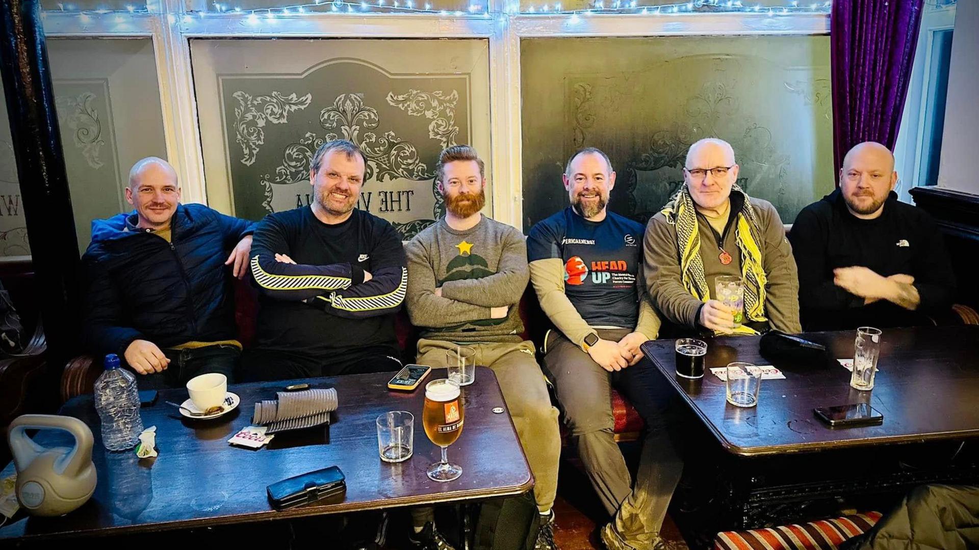 The six veterans sitting in a pub on completion of their walk. They are sitting in a row facing the camera. In front of them are two wooden tables with drinks.