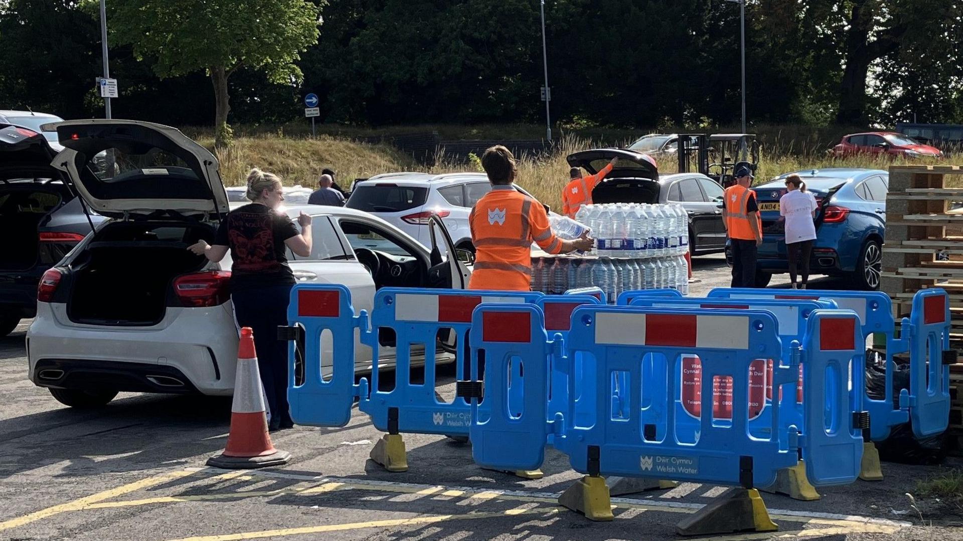 Bottled water station in Flintshire. Cars are parked beside the stations which have packs of bottled water. People are standing by their cars while volunteers in orange neon vests help with carrying the packs. 

