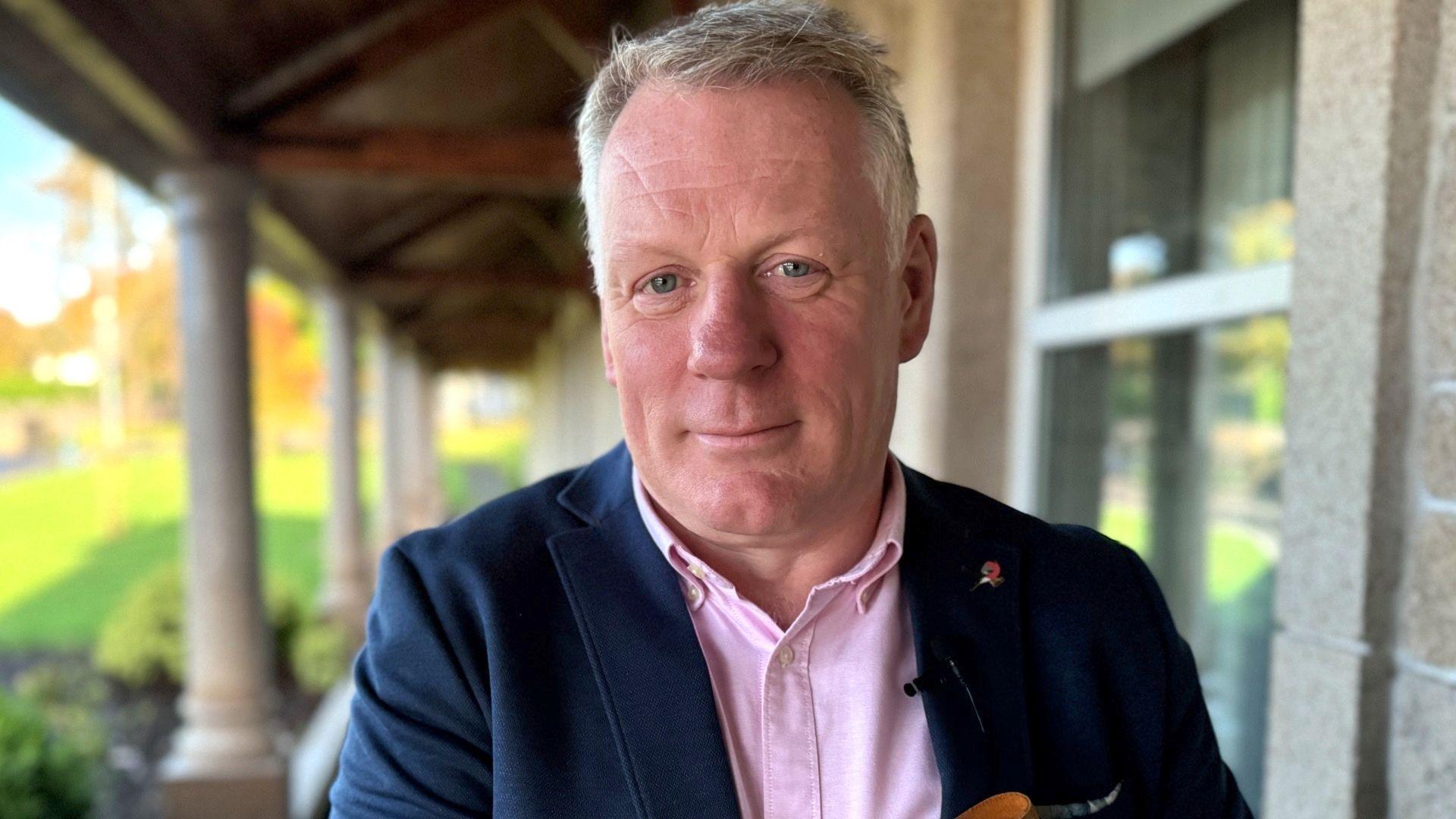 Johnny Finch looking straight to camera, wearing a pink shirt and navy blue jacket. He has grey hair.