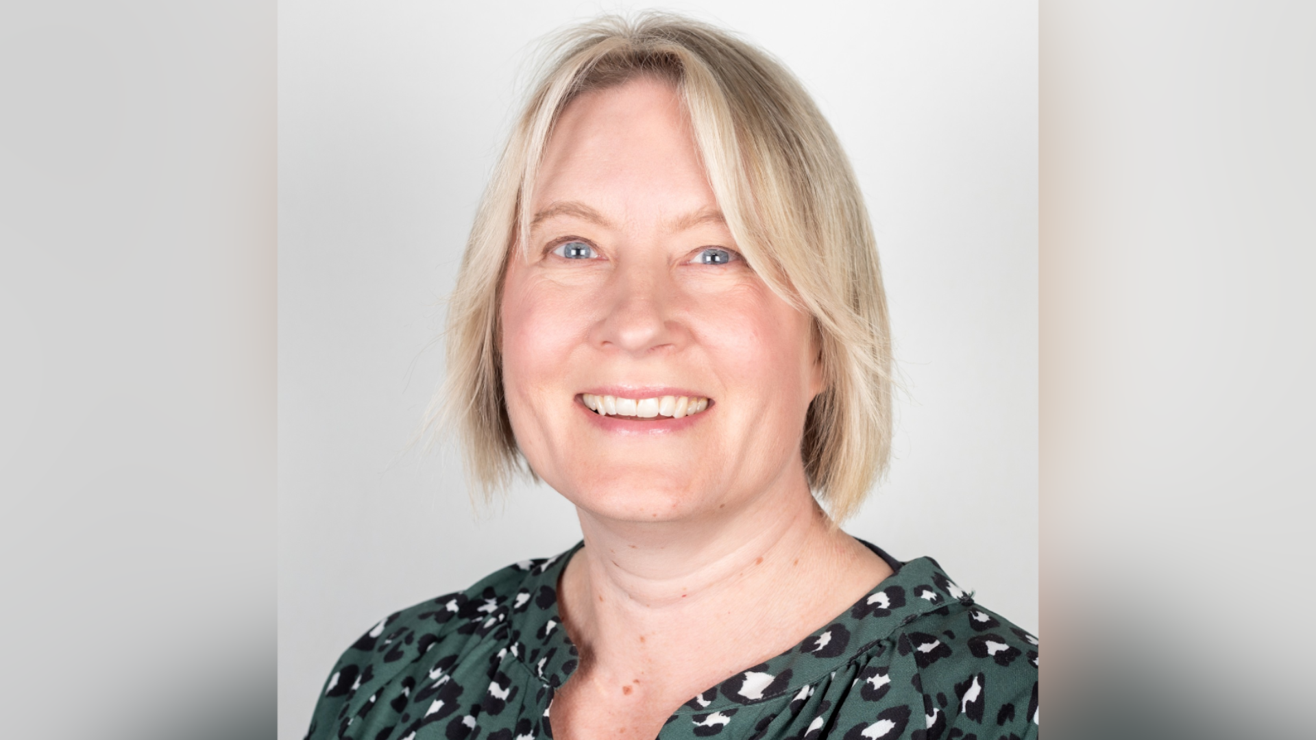 Hayley Douglas is smiling at the camera. She has blonde hair in bob style, blue eyes and is wearing a green, black and white dotted blouse. Behind her is a plain white background.