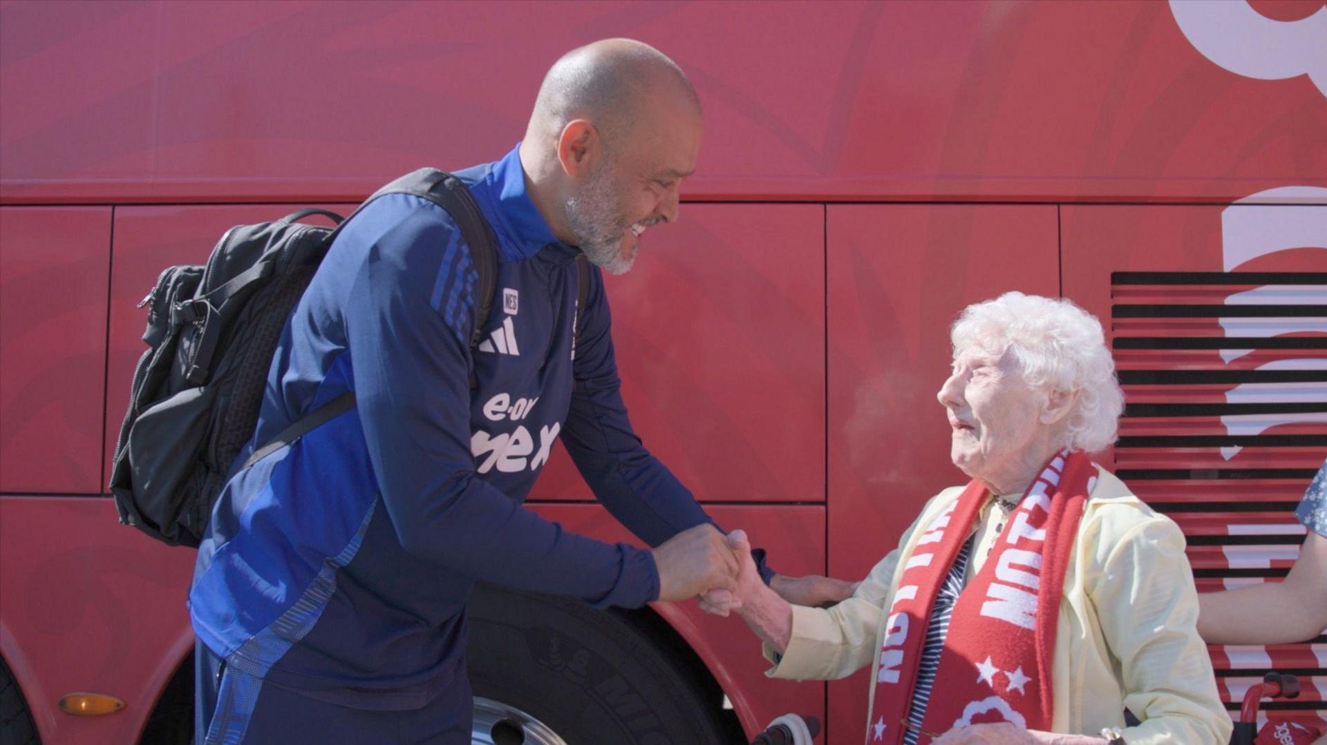 A man in a tracksuit smiles and shakes the hand of an older lady in red Forest scarf