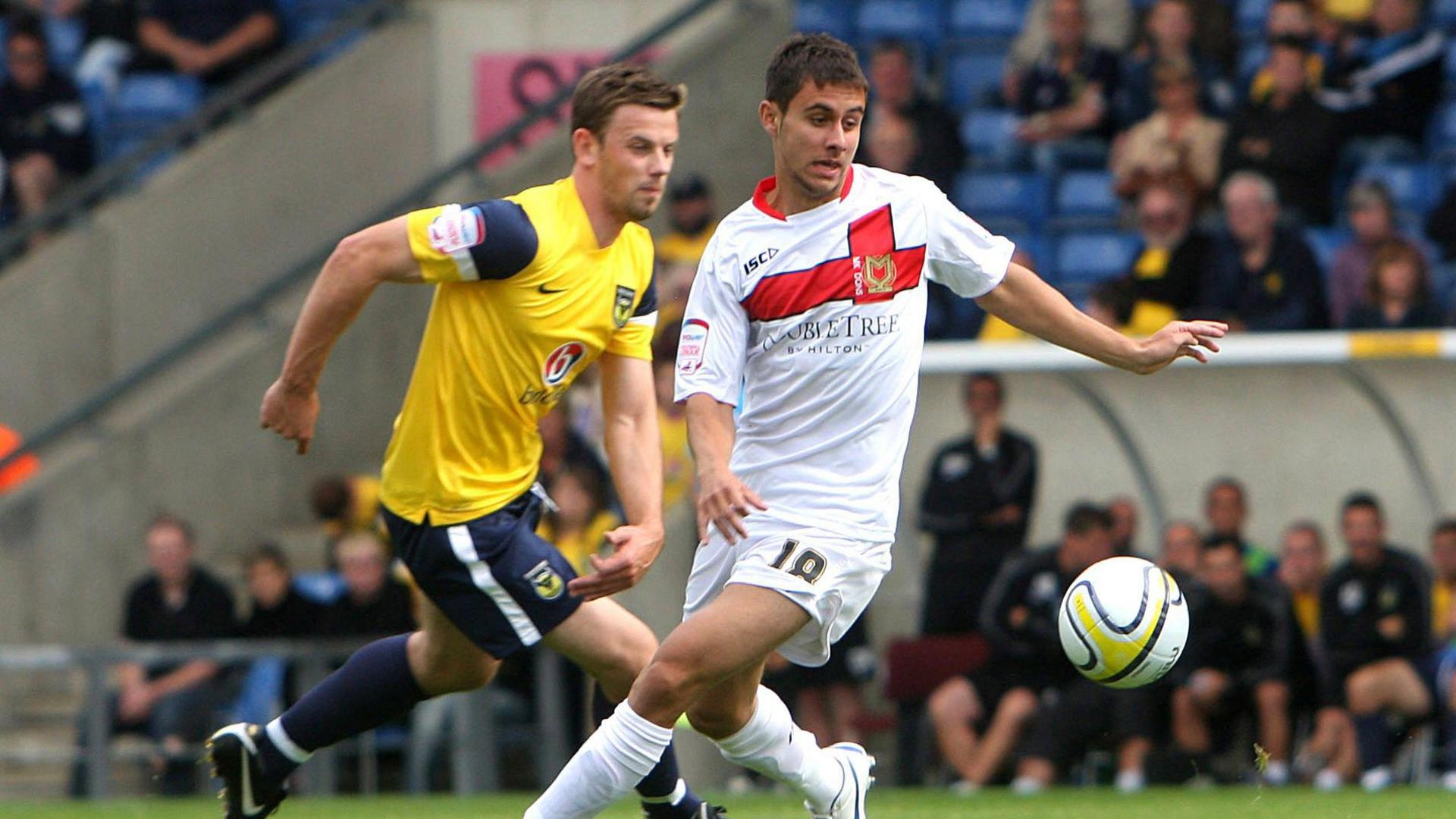 Action shot of George Baldock in a white kit controlling a mid-air ball under pressure from another player wearing a yellow and blue kit. 