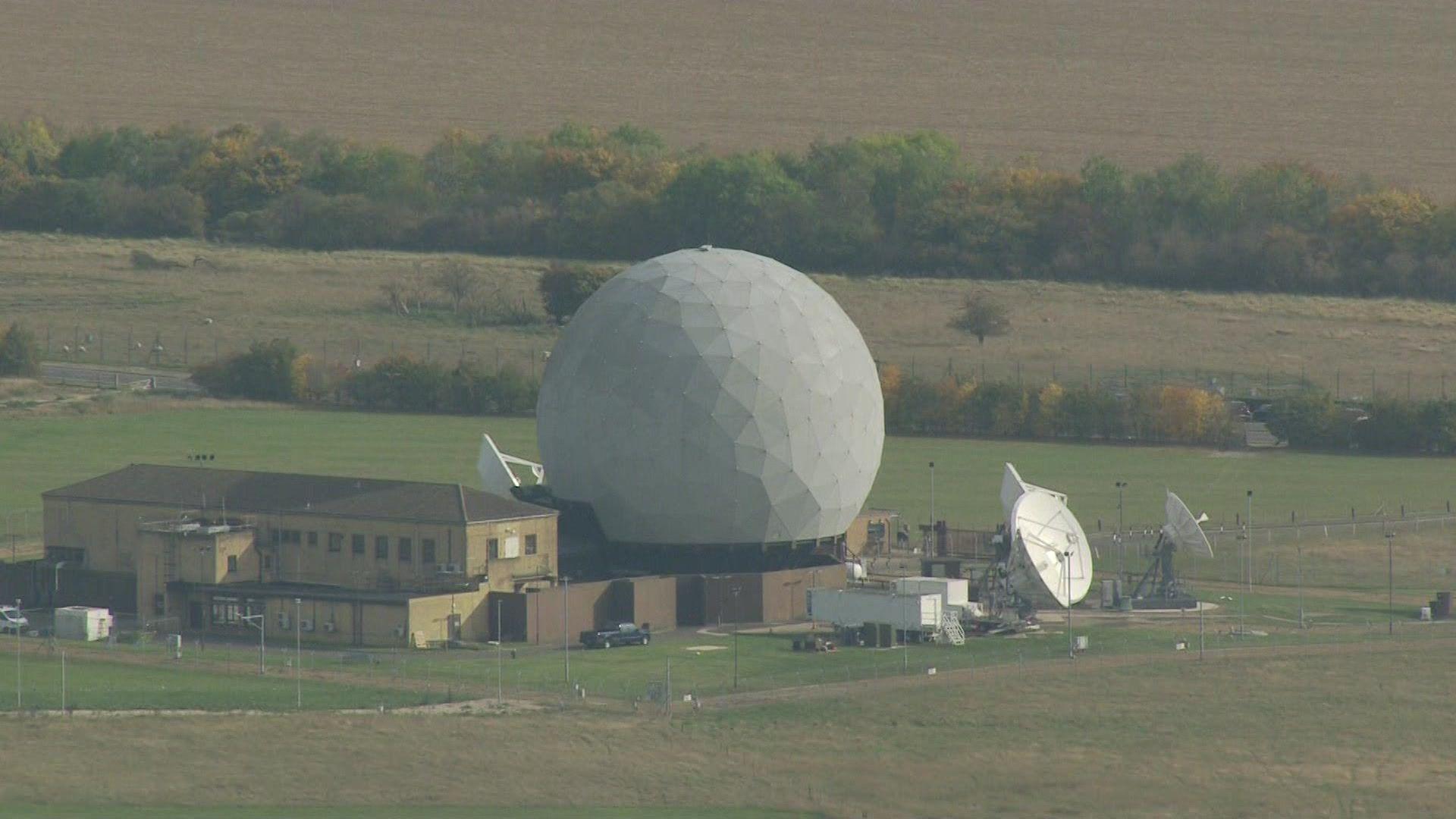 An picture from the air of a large golf ball like structure located on top of a single storey building. About four satellites are dotted around it. There is also a large two-storey building next to it. It is surrounded by fields.
