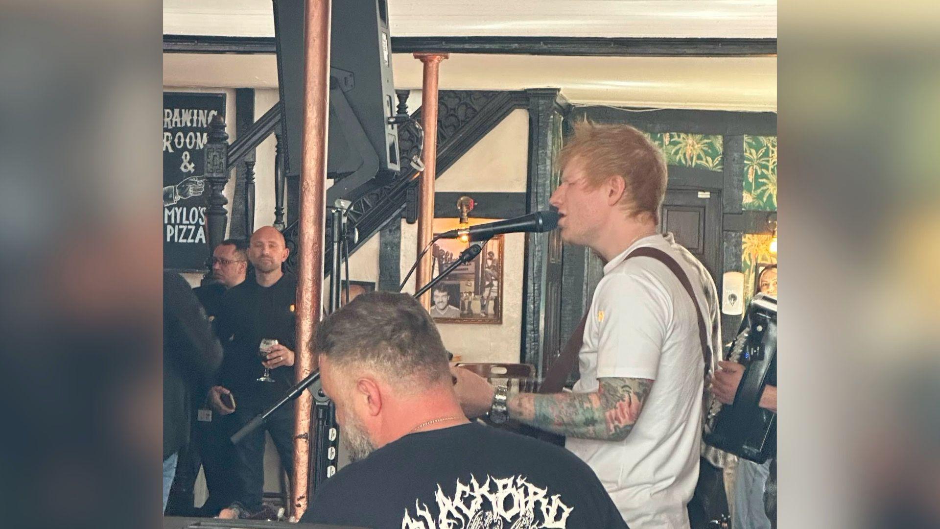 Ed Sheeran in a pub, playing his guitar and singing. Other musicians members can be seen. people mill about holding drinks.