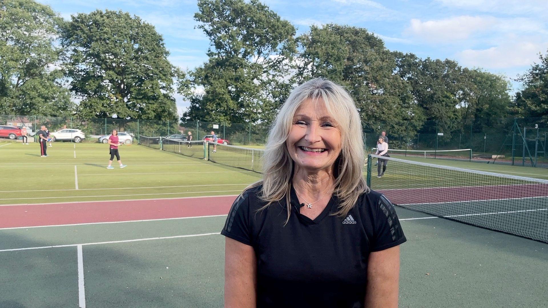 Gail Bromley standing on a tennis court with people playing behind her