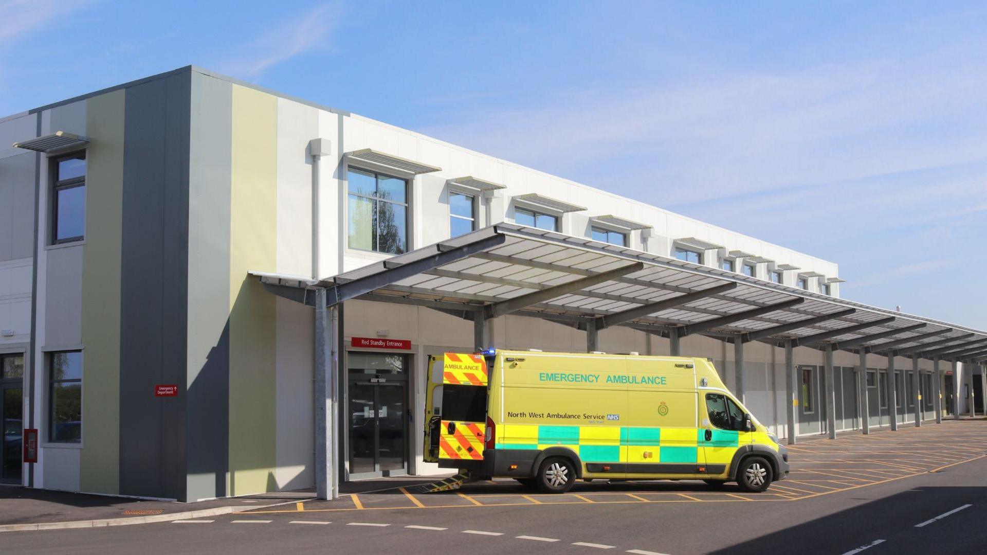 An ambulance is parked outside a modern-looking two-storey building with a glass canopy on the side of it. The ambulance's back doors are open.